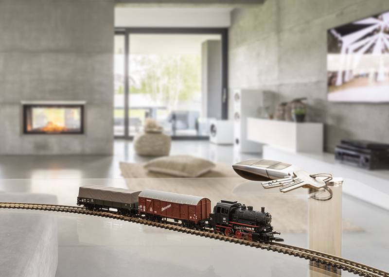 A miniature railway travels along a track through a modern living room. A fireplace and furniture can be seen in the background.
