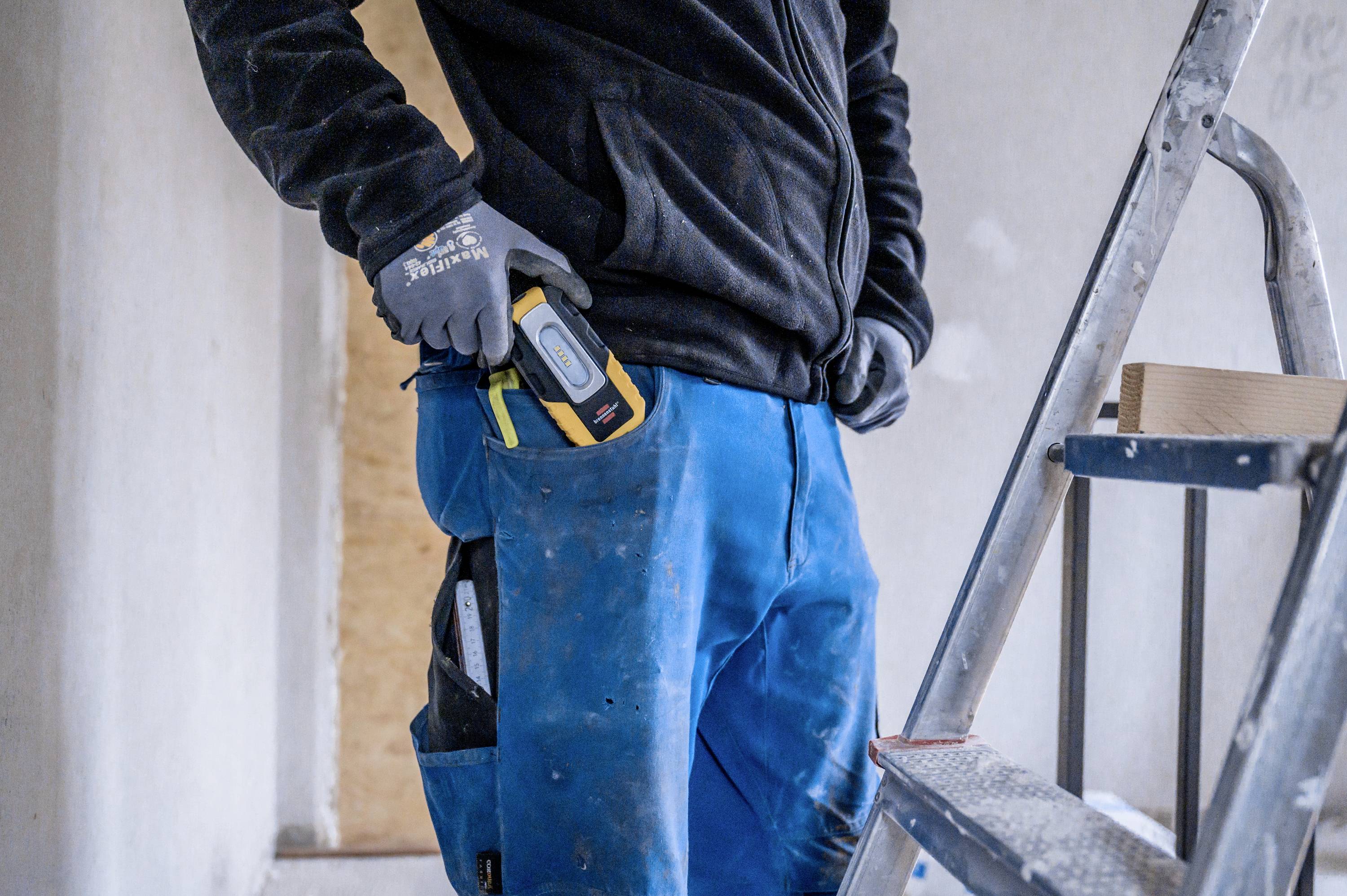 Tradesman on a ladder holding a measuring device, wearing blue work trousers and a black jacket, standing in an unfinished room.