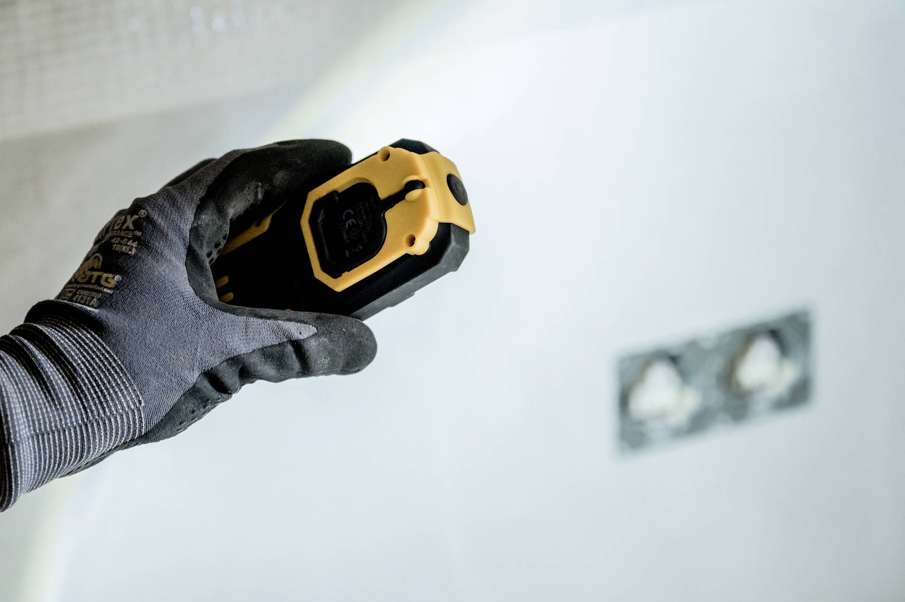 A person wearing a work glove is holding a yellow and black tool against a wall. A partially obscured socket is visible in the background.