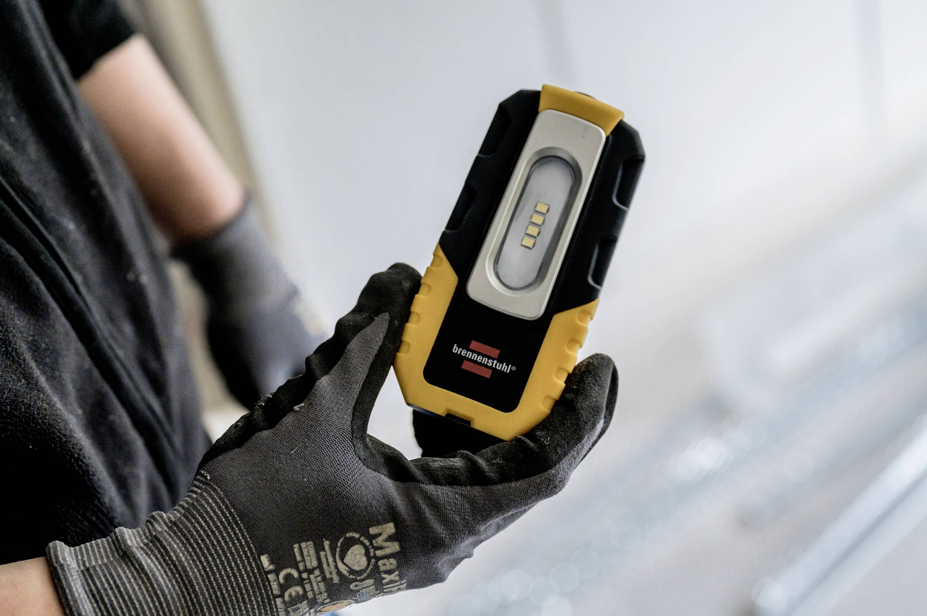A person is holding a work light with a yellow and black casing and a visible LED light bar in a gloved hand.
