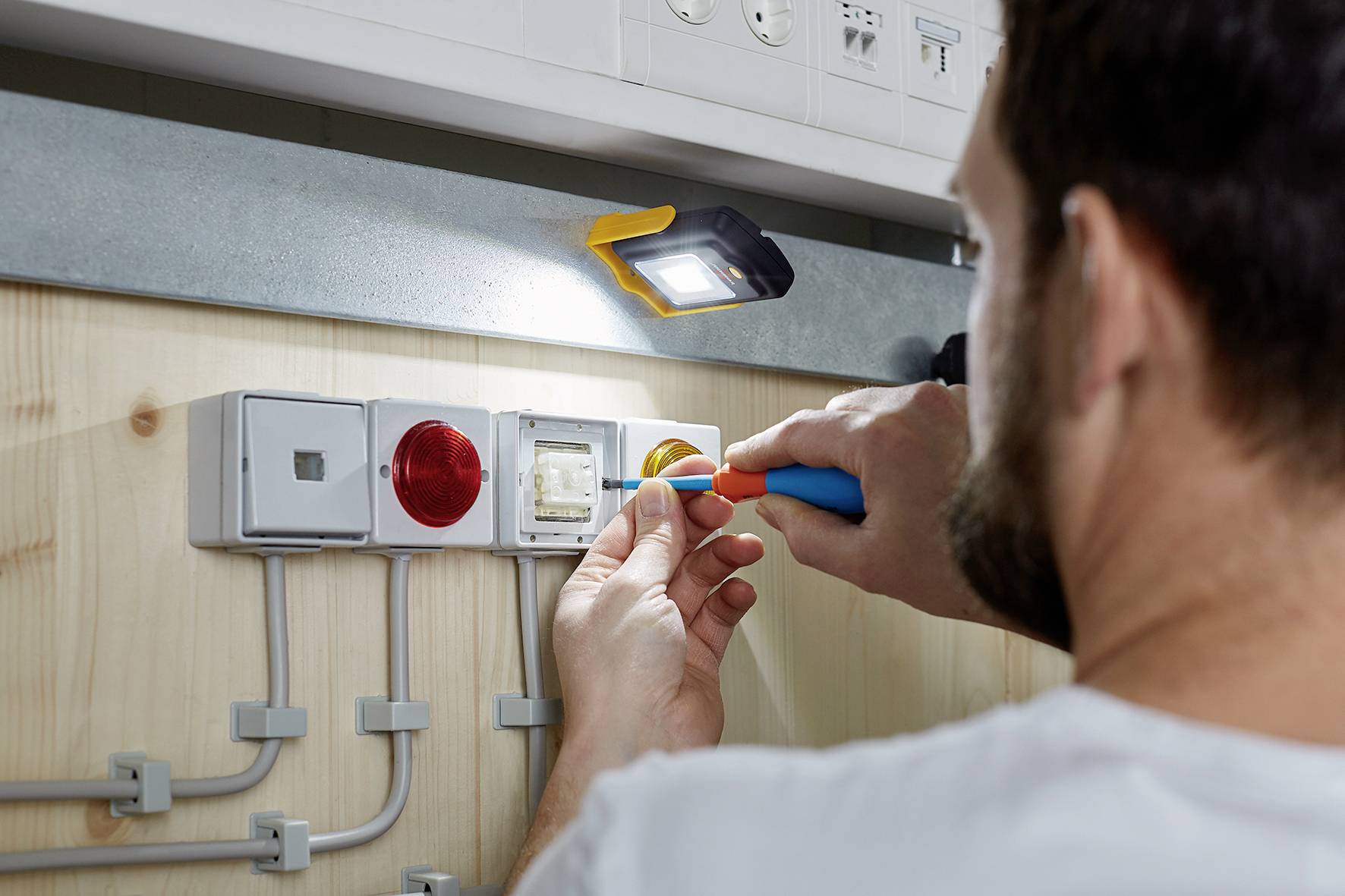 An electrician is working on a socket in a wooden panel, with a lamp illuminating the area.