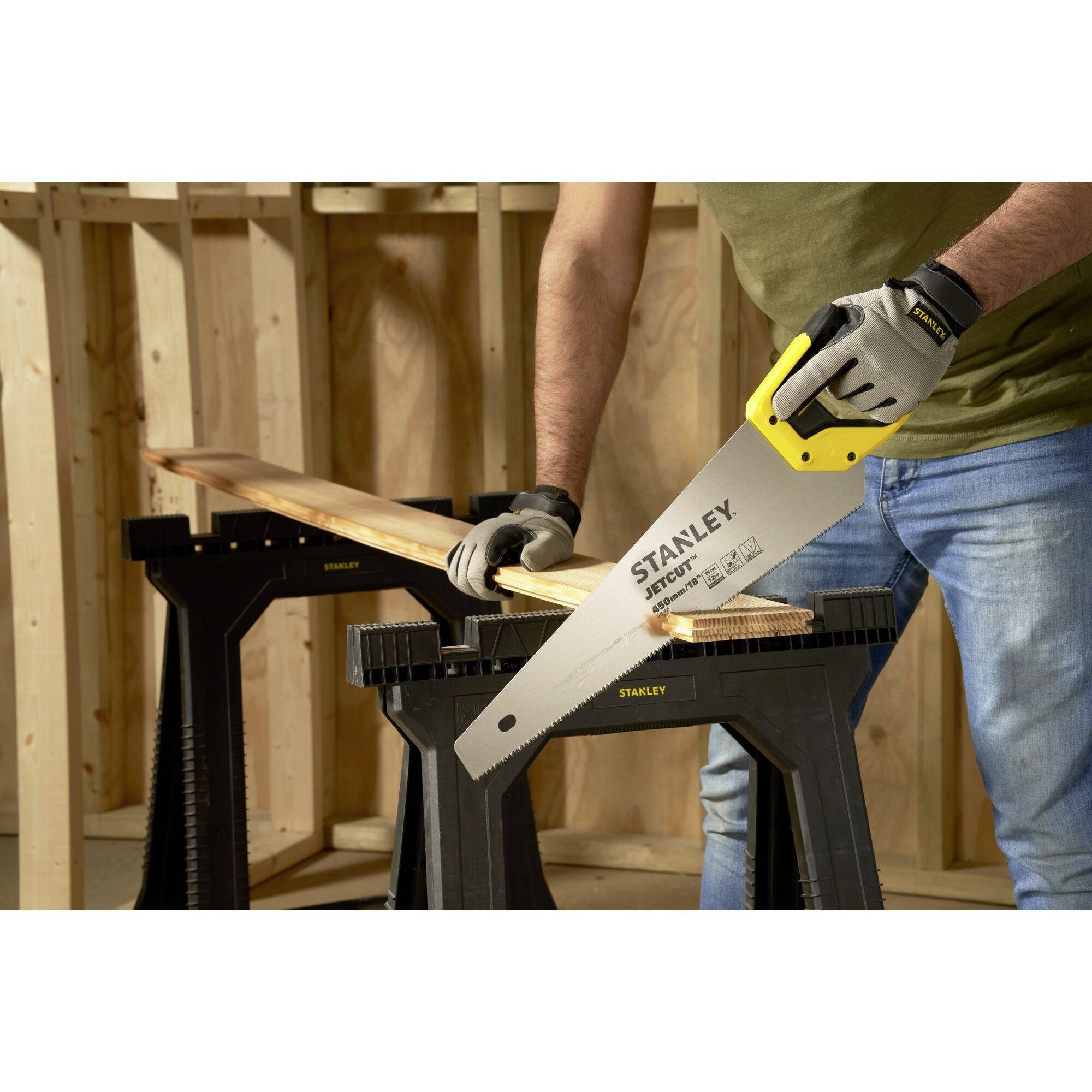 A man is sawing a wooden board with a handsaw, which is resting on two sawhorses. He is wearing work gloves and is standing in a workshop.