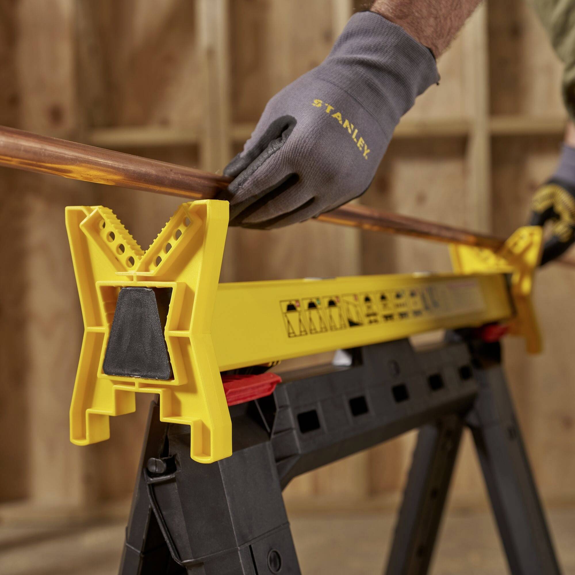 A person wearing gloves is holding a copper pipe on a yellow sawing horse in a workshop.
