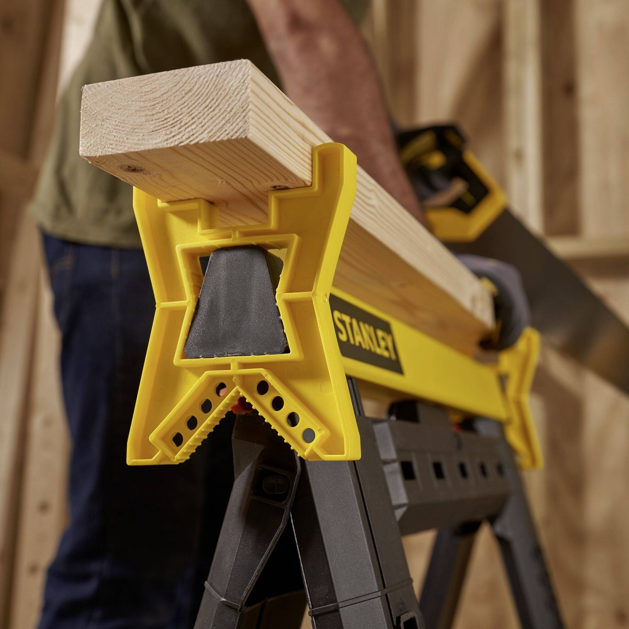 A person is sawing a wooden board with a handsaw, which is resting on a yellow workbench with a sawing support.