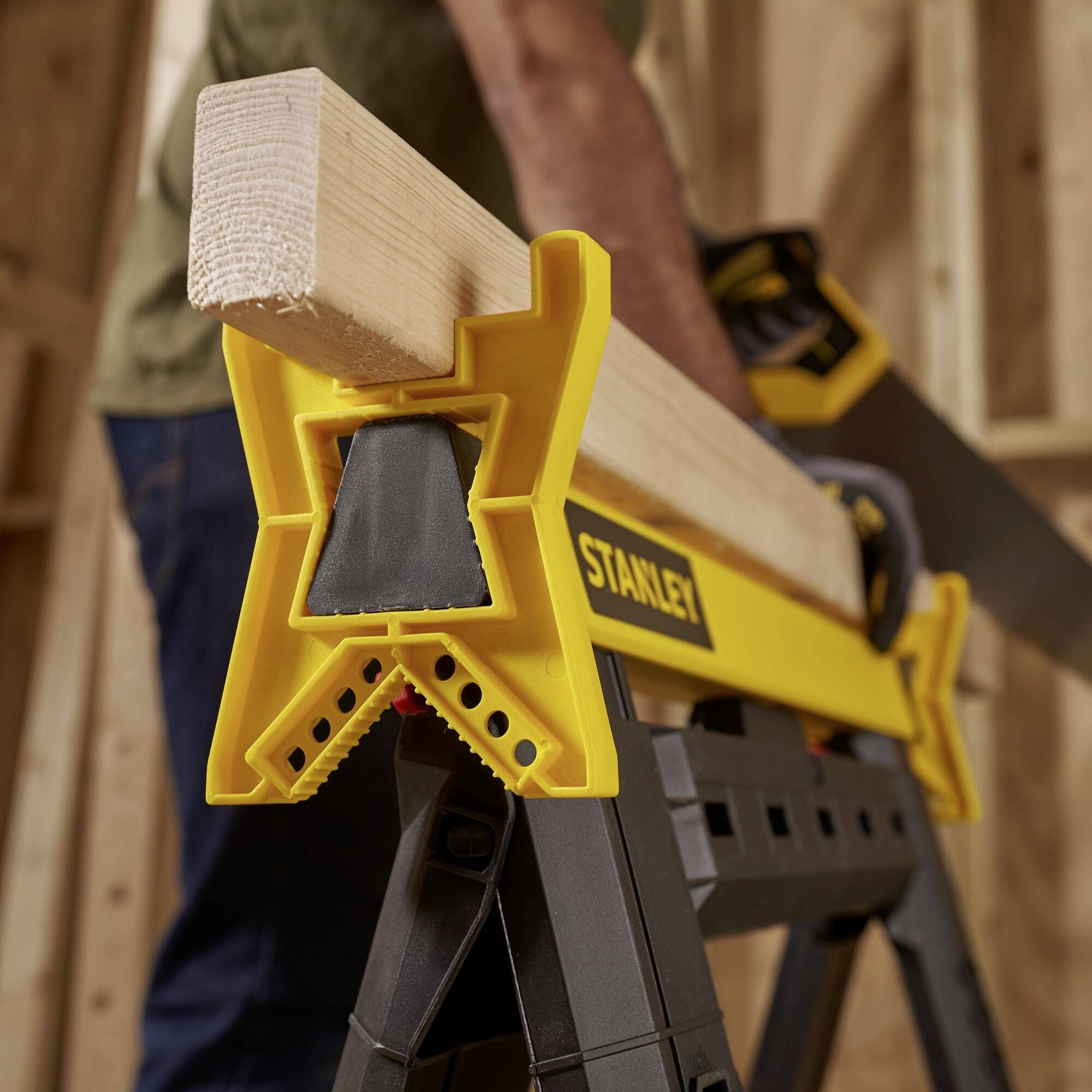 A yellow Stanley sawhorse is holding a wooden beam in a workshop environment. A person is sawing the beam.