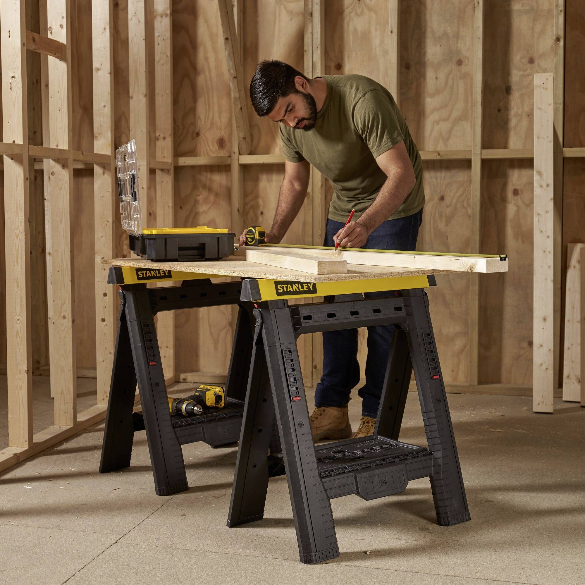 A man is working with a wooden board on a workbench in an unfinished room. Tools and wooden beams are positioned beside him.