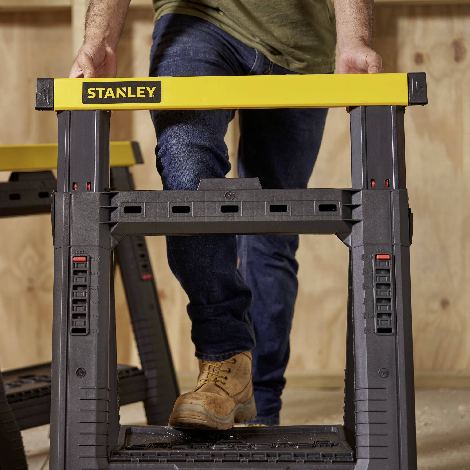 A person wearing jeans and boots is standing on a yellow Stanley tool stand in a workshop environment.