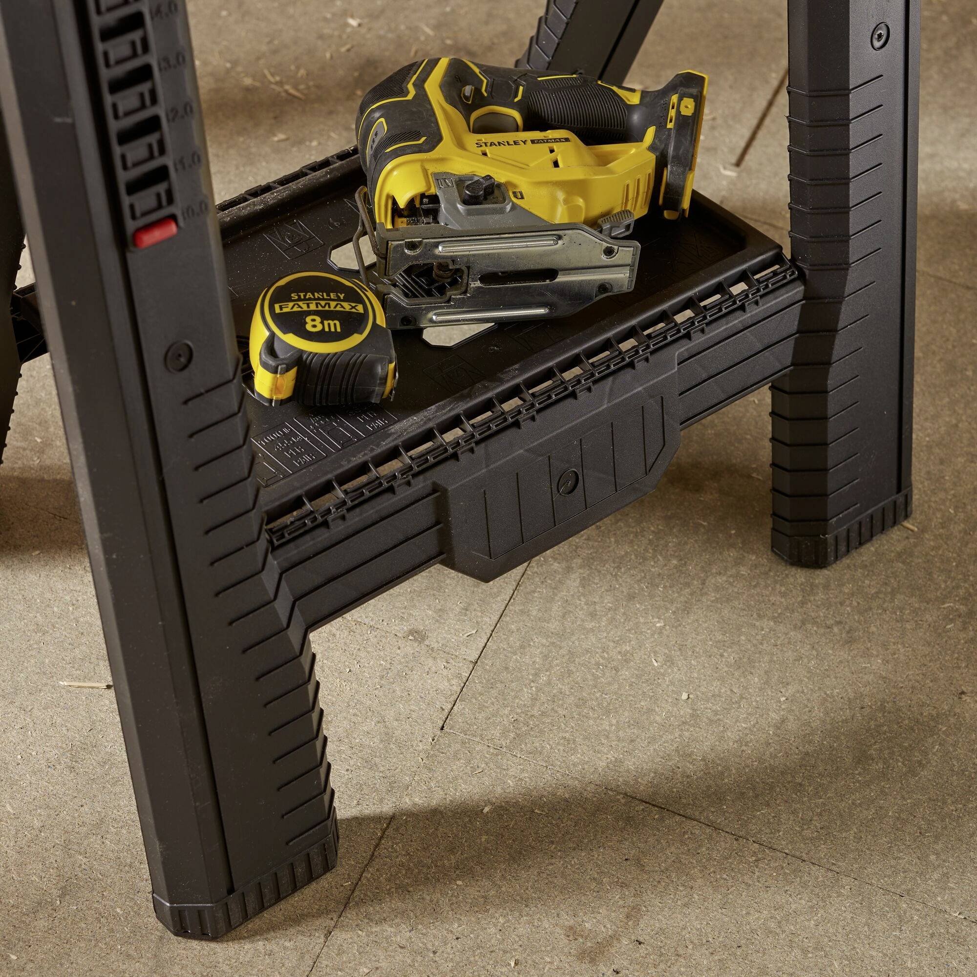 A yellow tape measure and electric tools lie on a black folding table. The background shows a wooden floor.