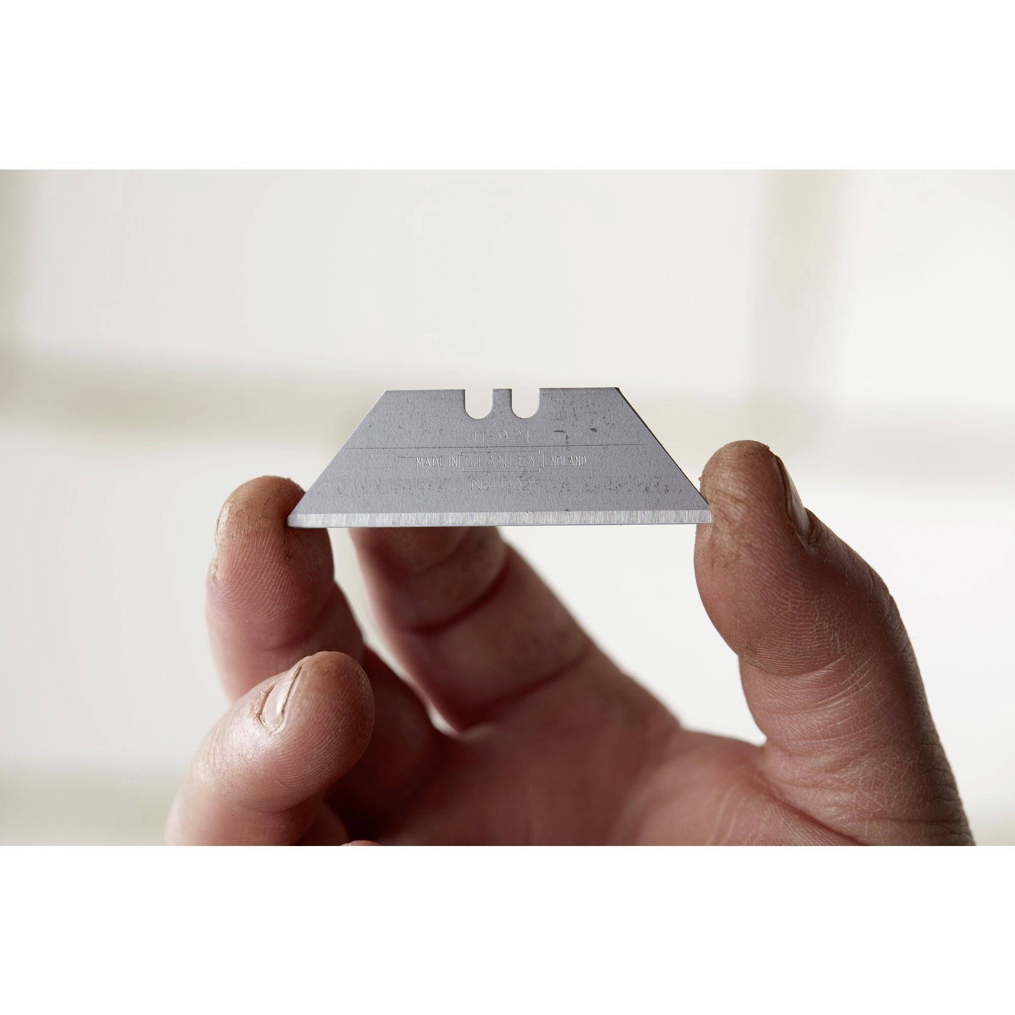 A hand holds a trapezoidal razor blade between thumb and forefinger, front view.