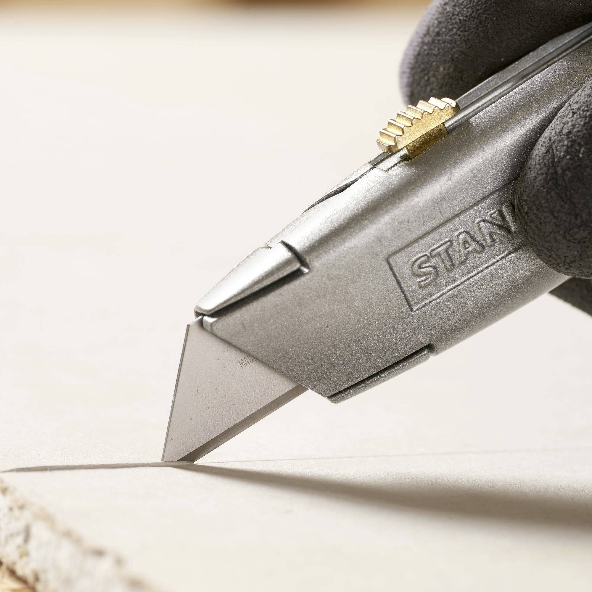 A silver carpet knife cuts through cardboard, held by a gloved hand.