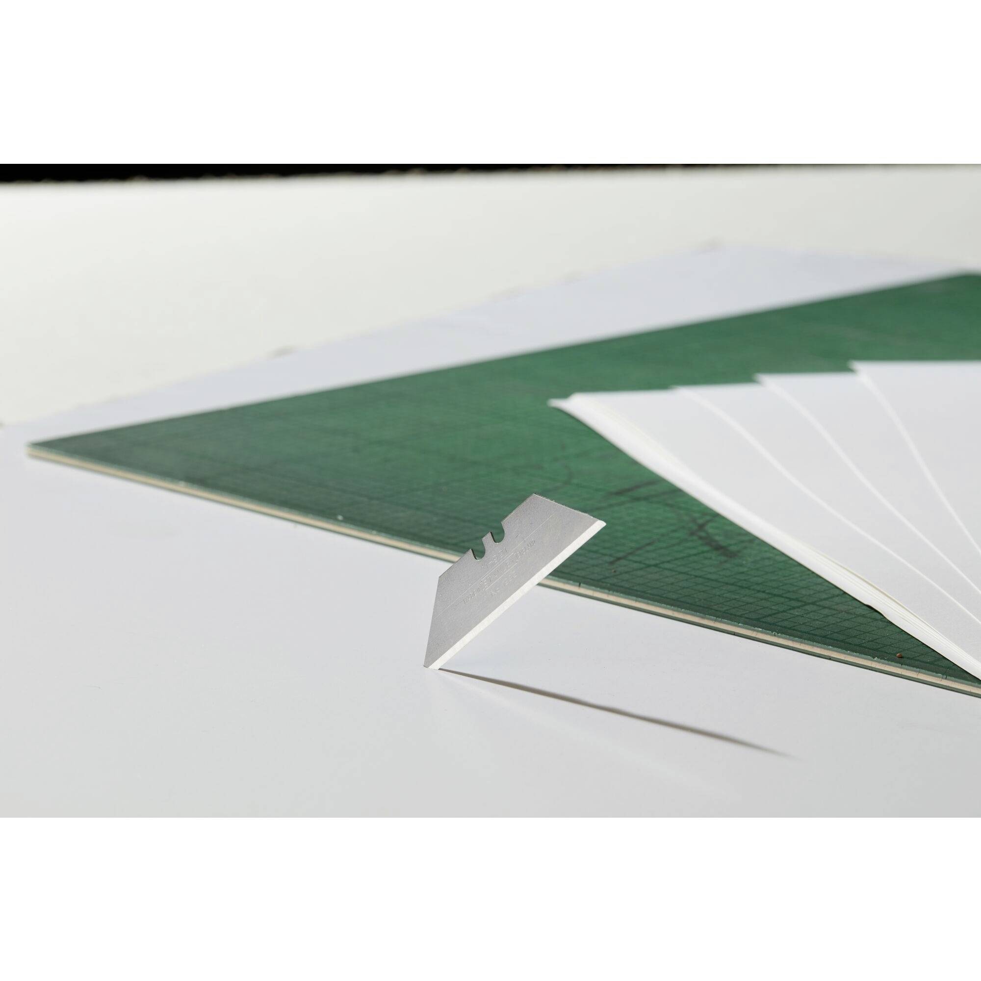 Paper lies on a green cutting mat. A craft knife is positioned diagonally on the paper, blade facing upwards.