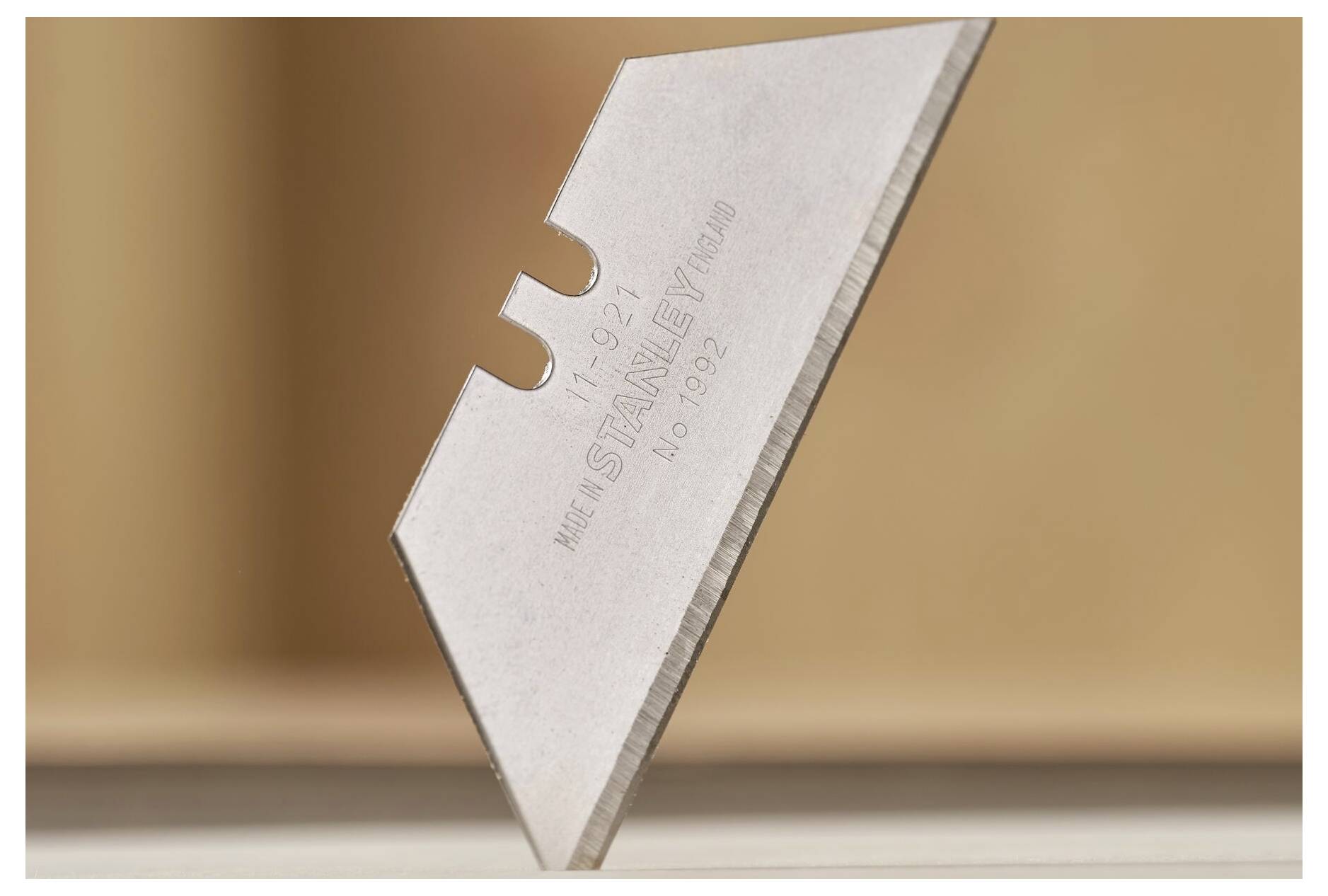 A metal blade piece, likely from a utility knife, stands on its point against a blurred background.