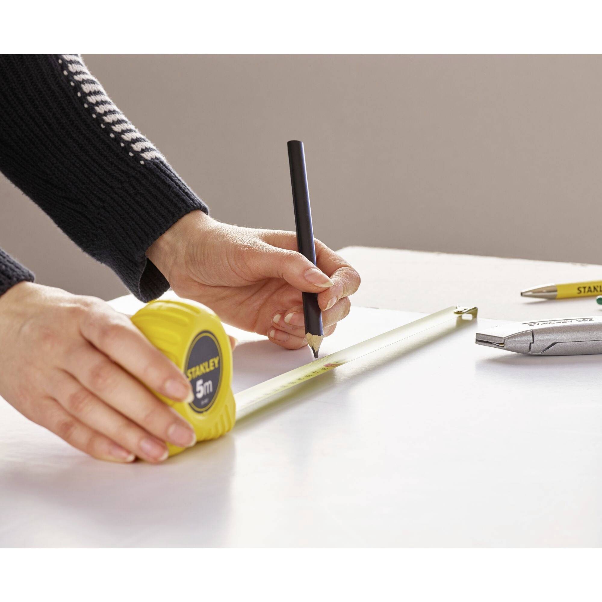 A person is holding a measuring tape, stretching it across a table and marking a point with a pen. Next to the measuring tape is a craft knife.