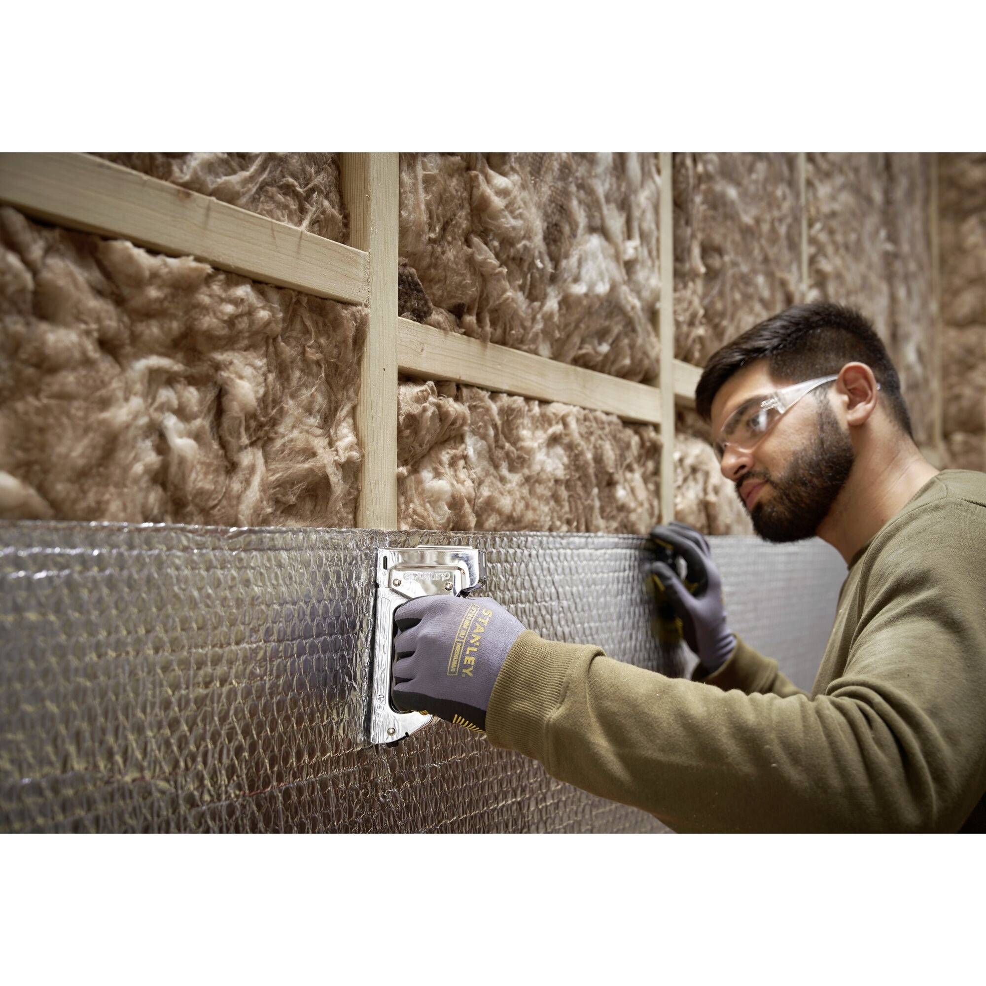 A man is securing thermal insulation to a wall, wearing safety glasses and gloves. Insulation material is visible in the background.