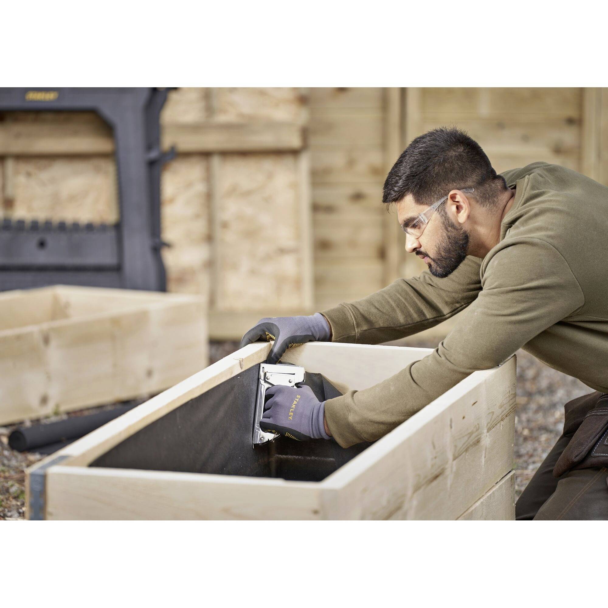 A man is sanding a wooden box using a grinding machine. He is wearing safety glasses and gloves. The background shows additional wooden planks.