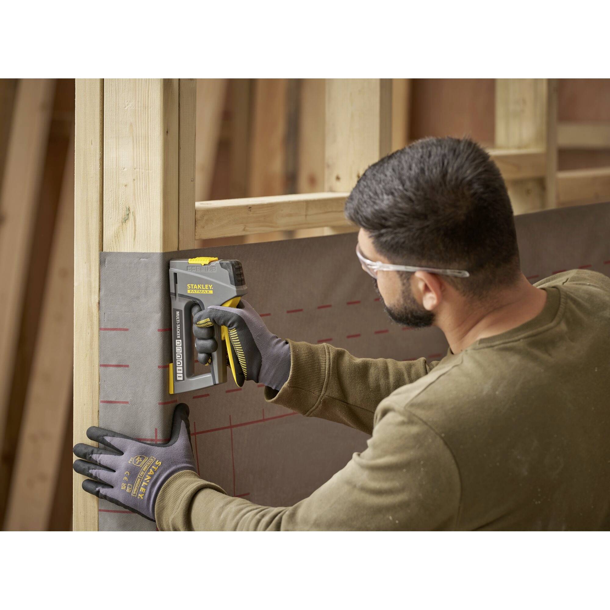 A man is attaching black and red material to wooden frames using a staple gun. He is wearing safety glasses and work gloves.