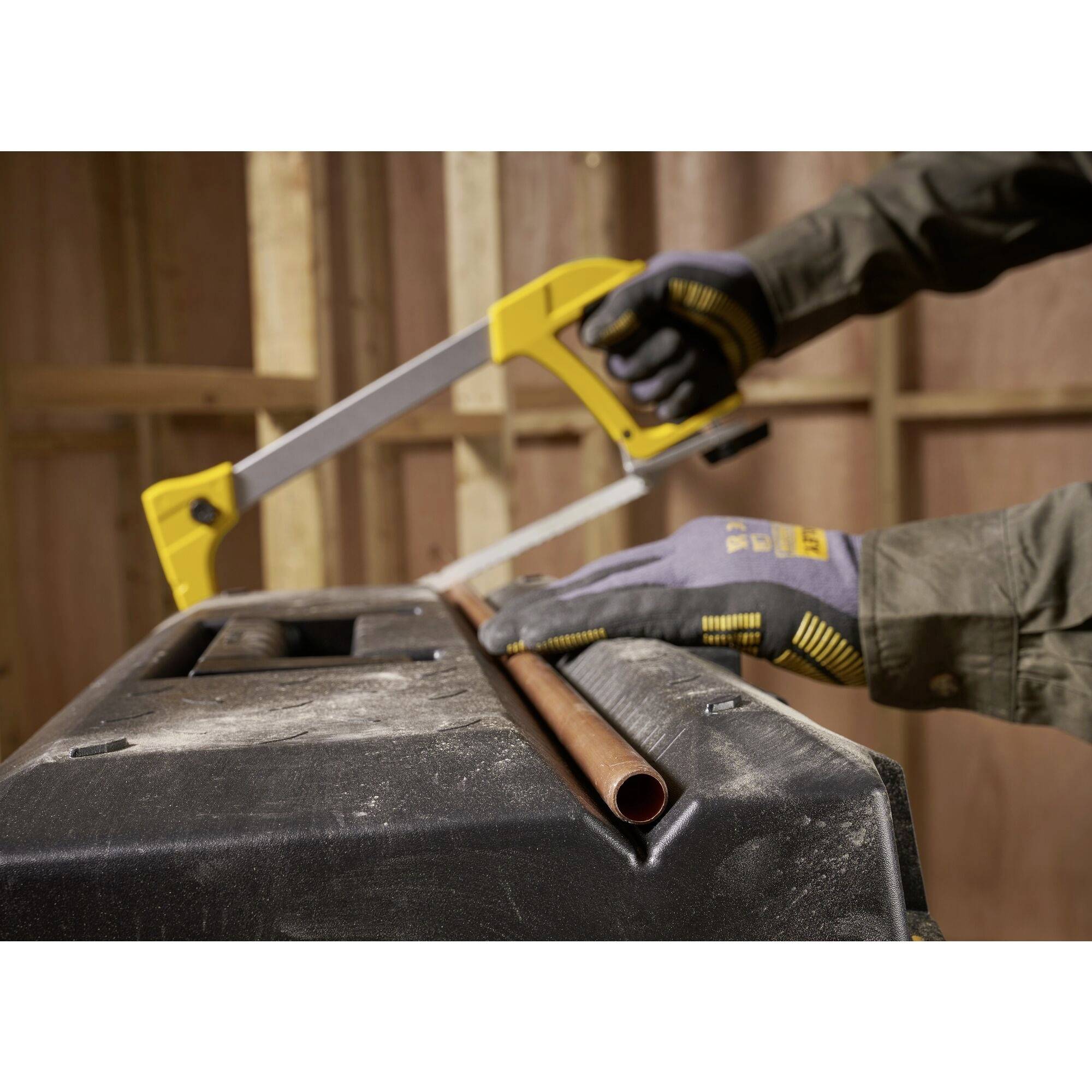 A person is sawing a copper pipe on a black work surface using a yellow handsaw. They are wearing work gloves and dark workwear.