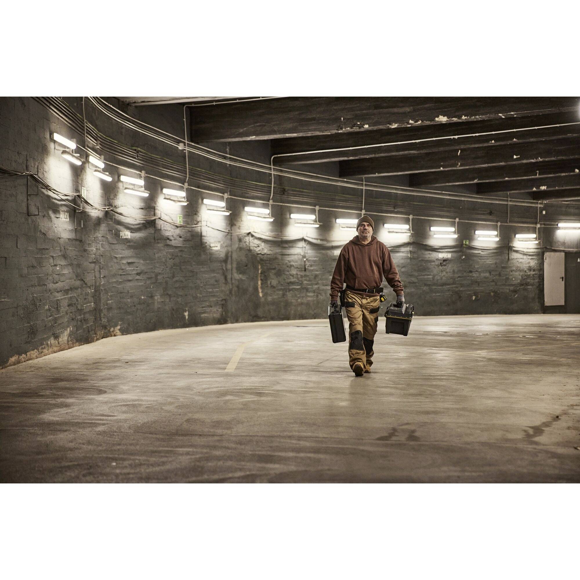 A person walks through an empty, illuminated car park, carrying two toolboxes. The environment is gloomy and industrial.