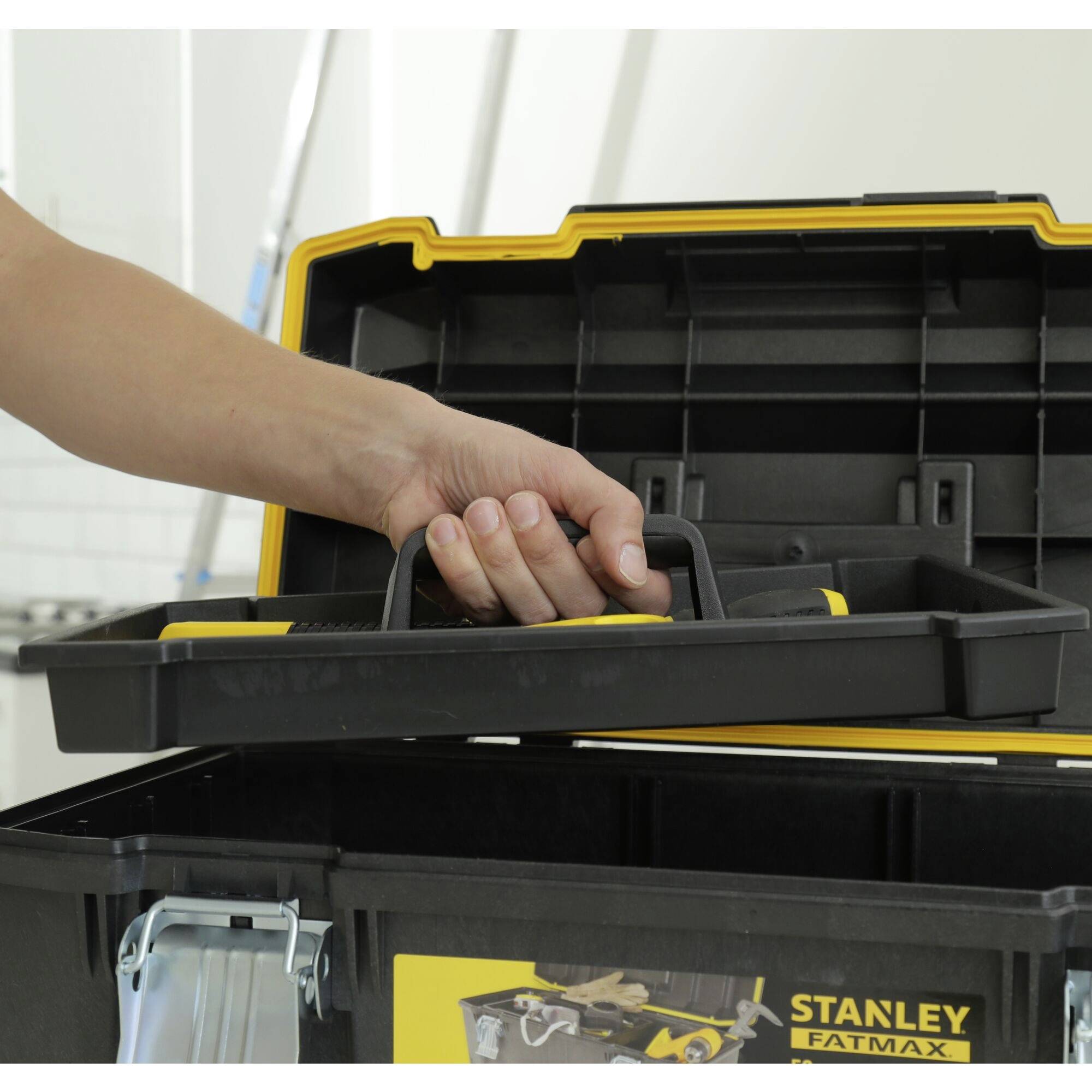 'Human hand reaches into an open, black toolbox with a yellow rim, which is standing in front of a light-coloured wall.'