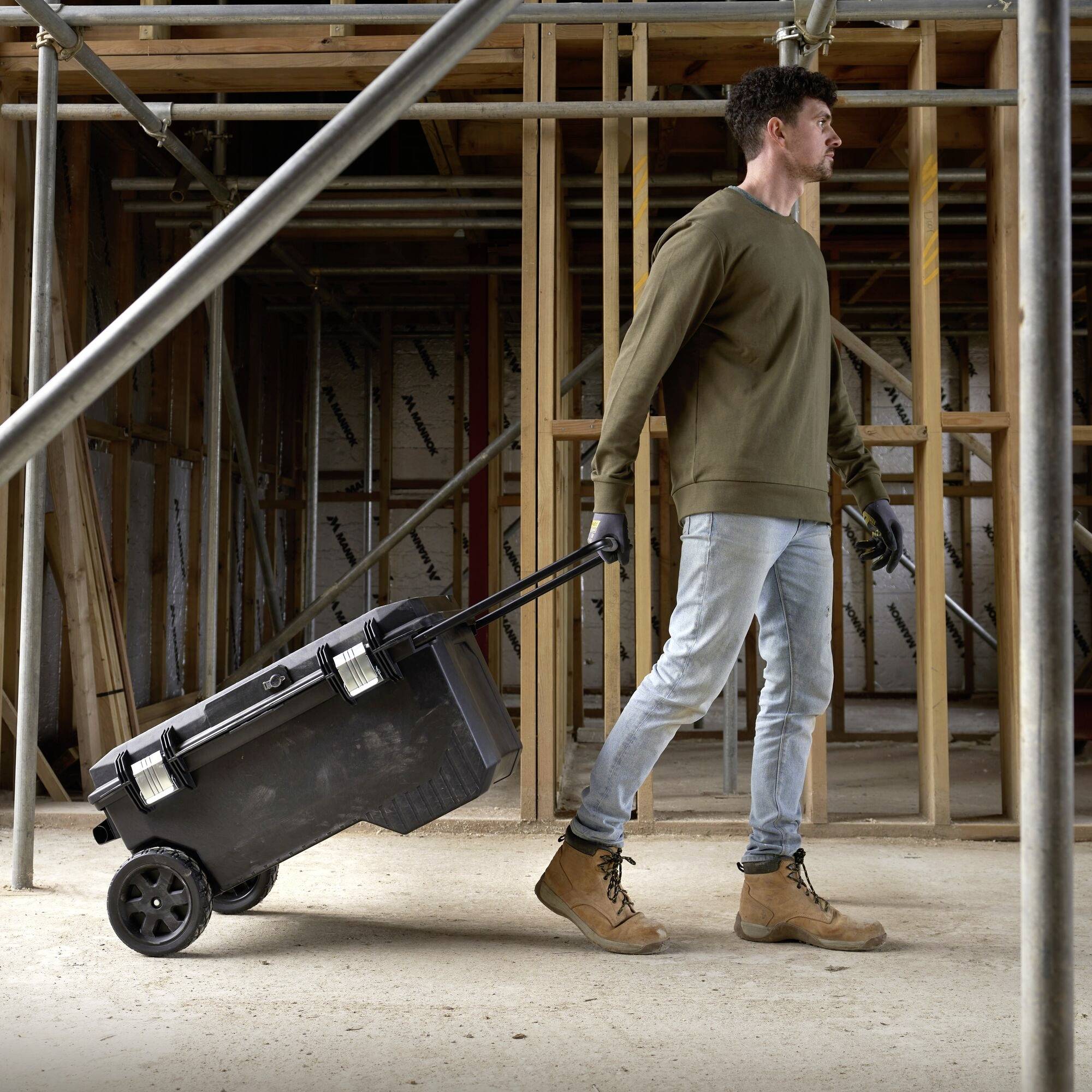 A man pulls a black toolcase on wheels through a construction site with wooden framing. He is wearing jeans and work boots.