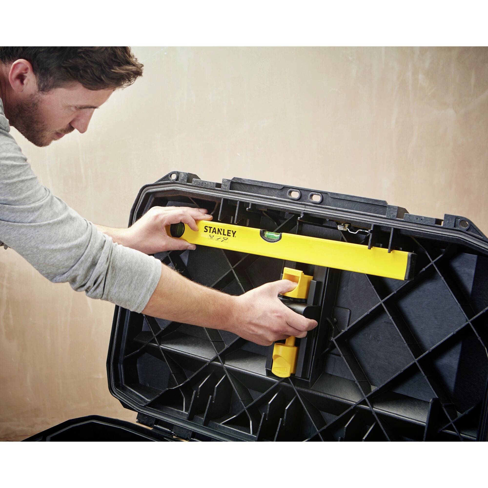 A man is placing a yellow spirit level into a black toolbox. He is wearing a grey long-sleeved shirt.