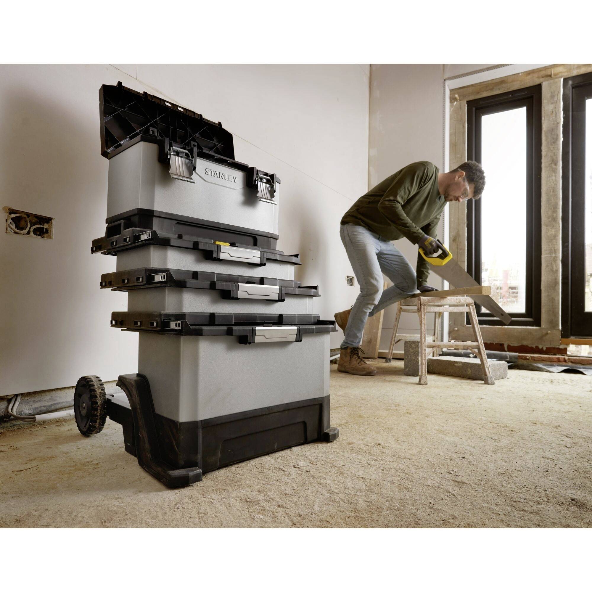 A man is working with a saw in an unfinished room; a stackable toolbox with wheels stands in the foreground.