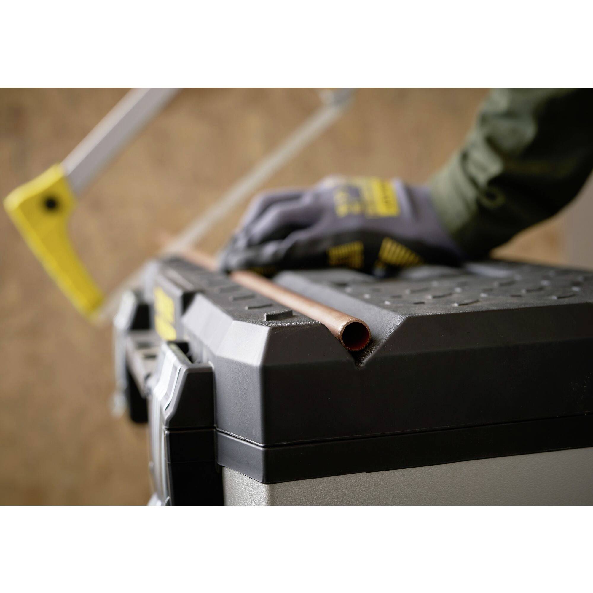 A person wearing gloves is sawing a copper pipe on a sturdy workbench. A saw can be seen in the background.
