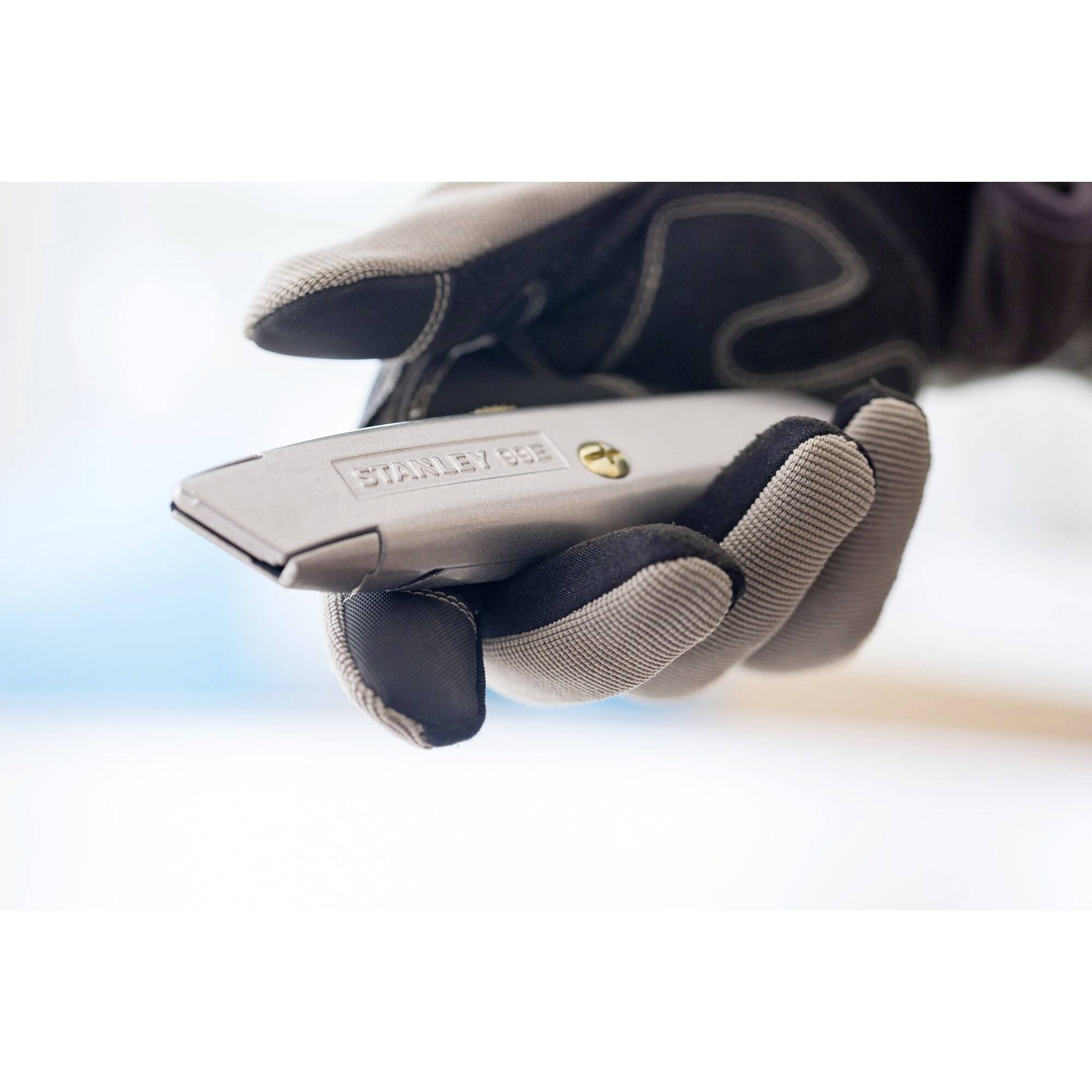 A hand wearing a work glove holds a grey Stanley Stanley knife, ready to cut.