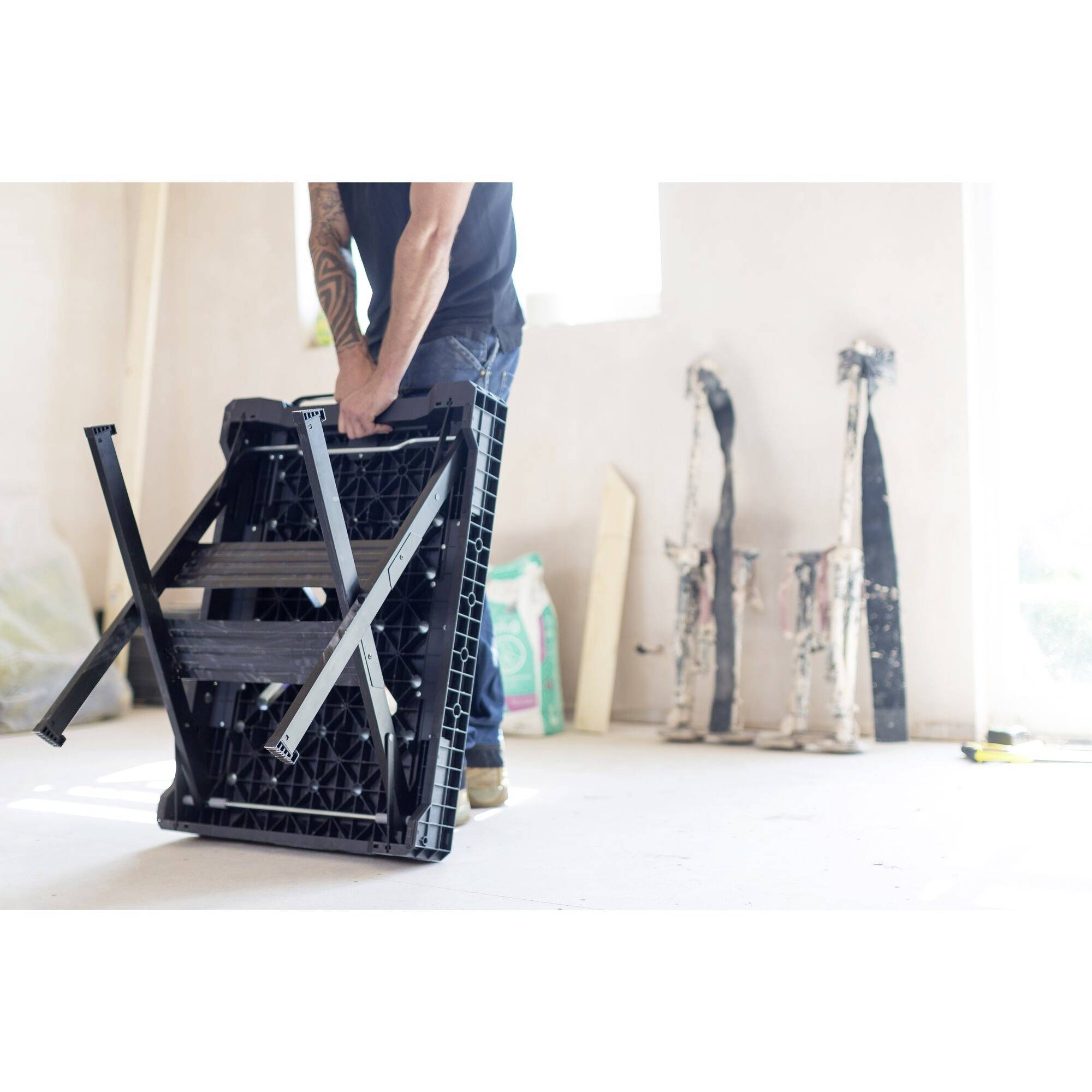 A person is dismantling a table in a bright room. In the background, folded painter's ladders and a sack are standing.