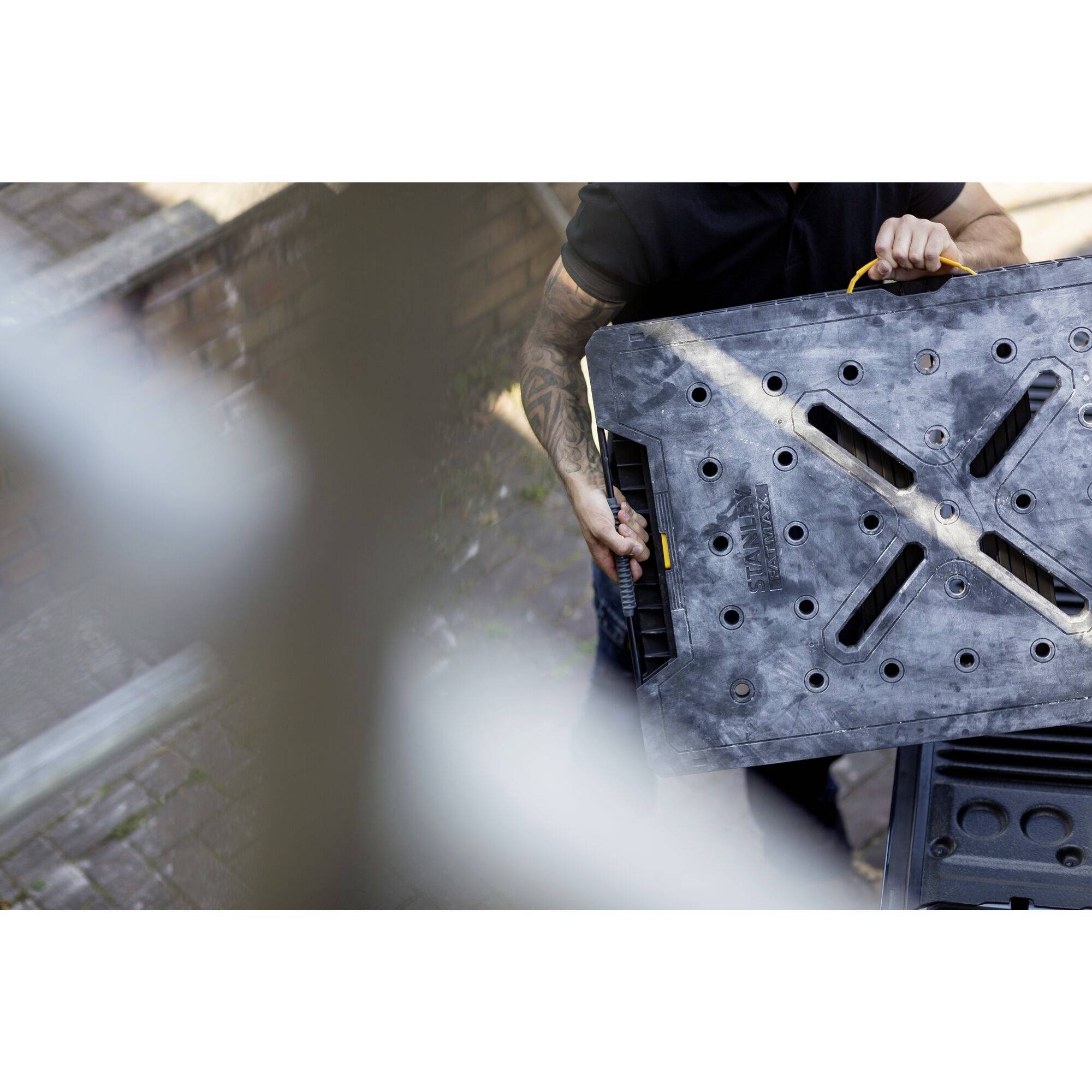 A person is carrying a large, flat, rectangular metal plate with several holes and handles. The scene is outdoors.