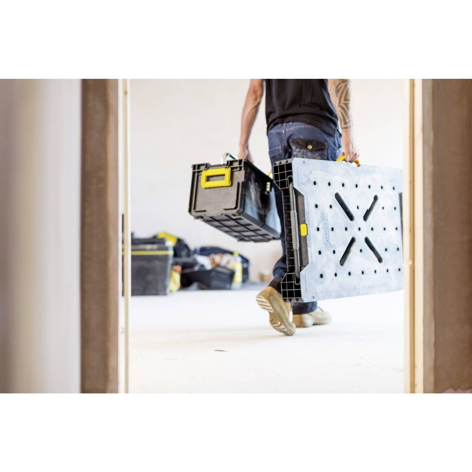 A person carrying a toolbox and a folded table frame is walking through an unfinished room. In the background, additional tools are lying around.