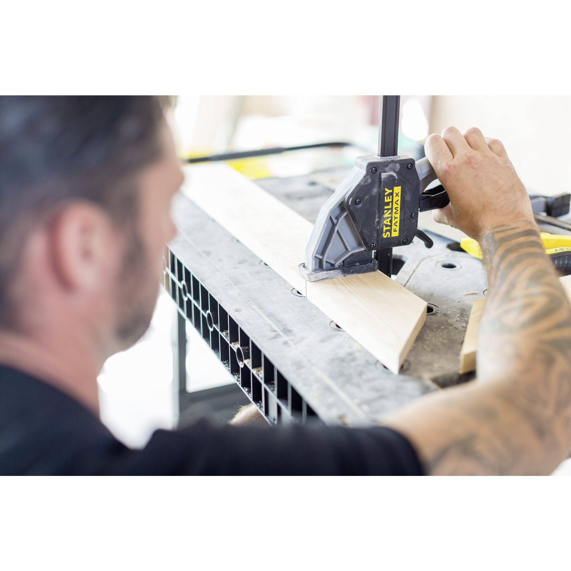 A man with tattooed arms uses a clamp to secure a piece of wood on a table. The focus is on craftmanship.