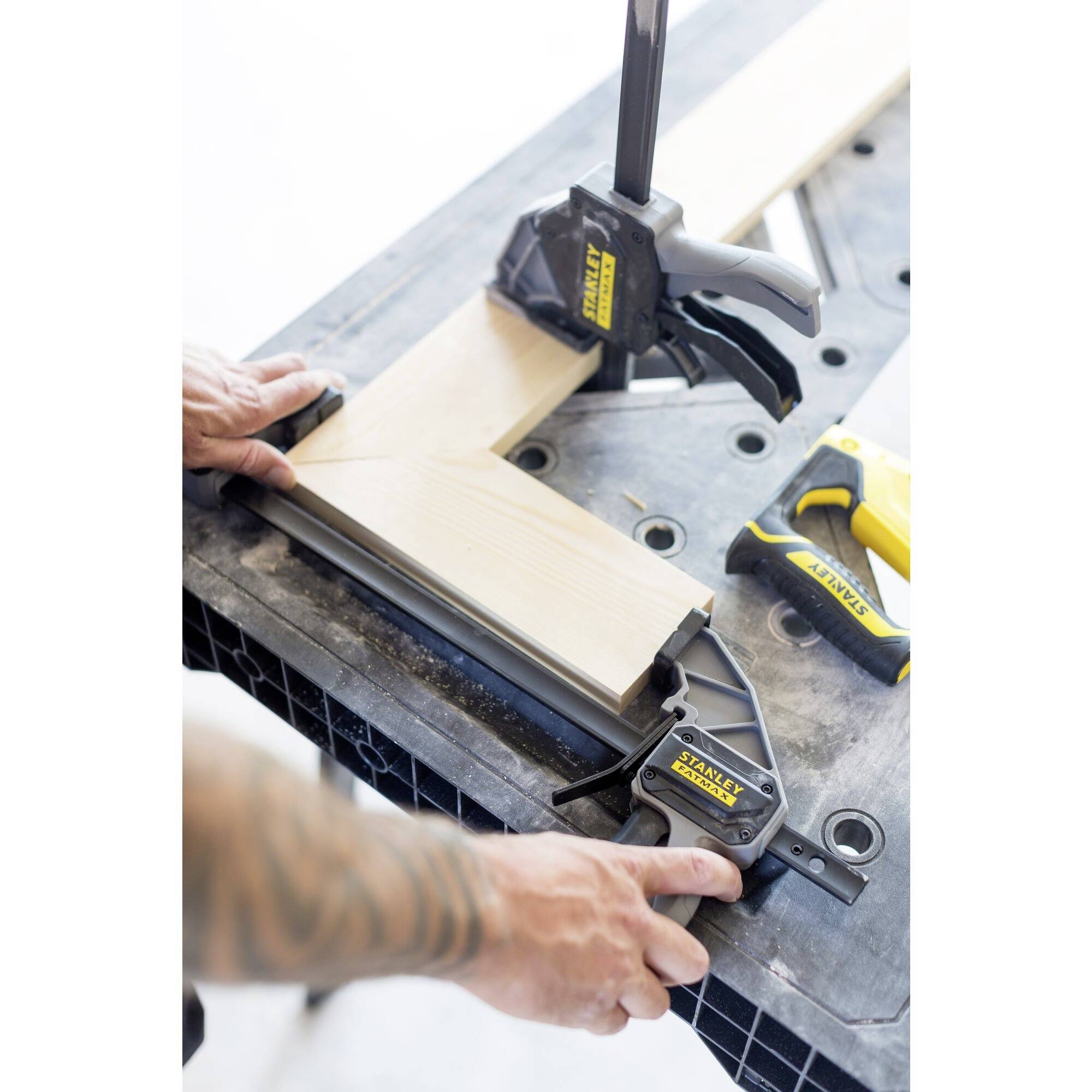 A person clamps two wooden boards at right angles to a table using G-clamps. A yellow handsaw lies beside them.