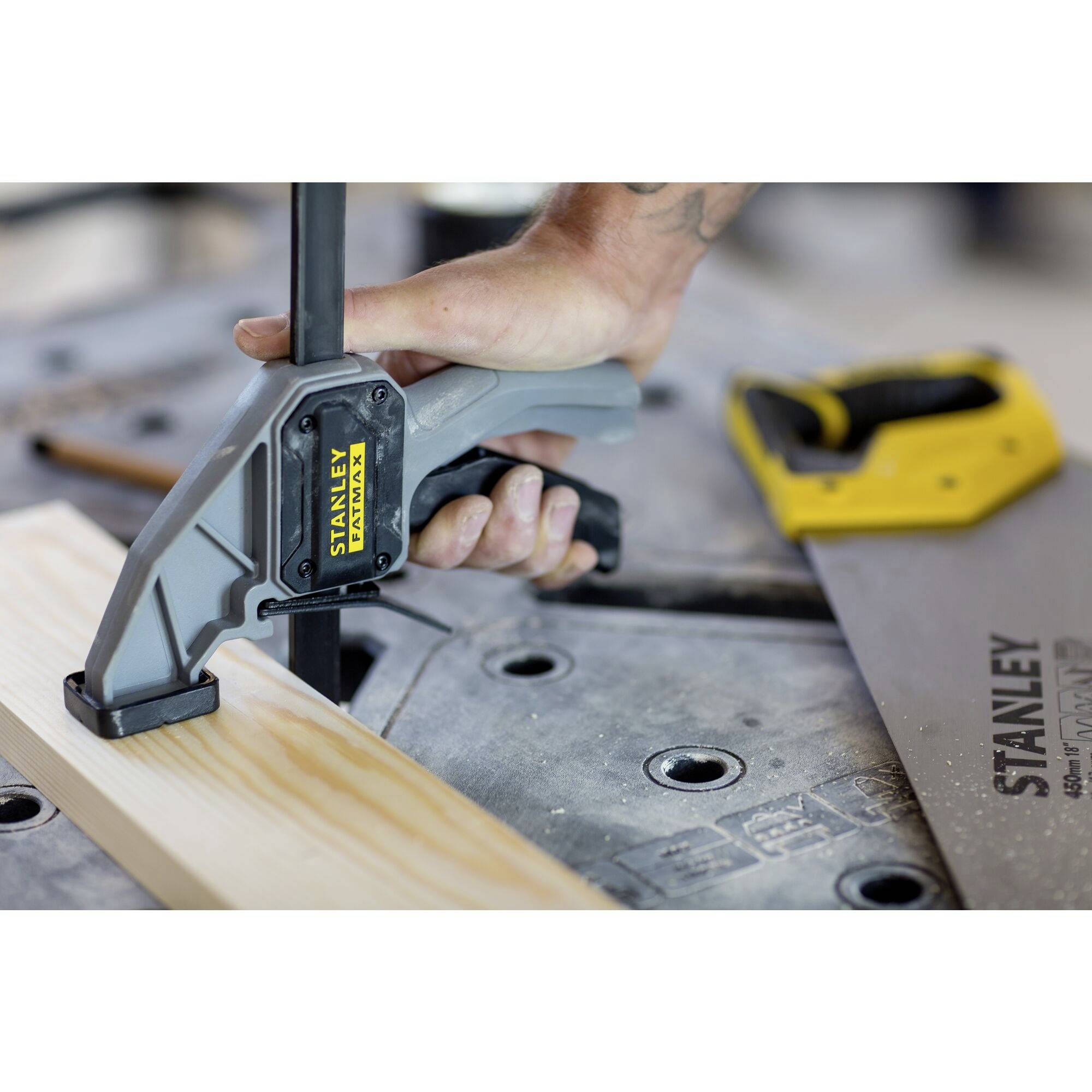 A hand is holding a Stanley clamp, which is securing a piece of wood on a workbench. Other tools are lying nearby.