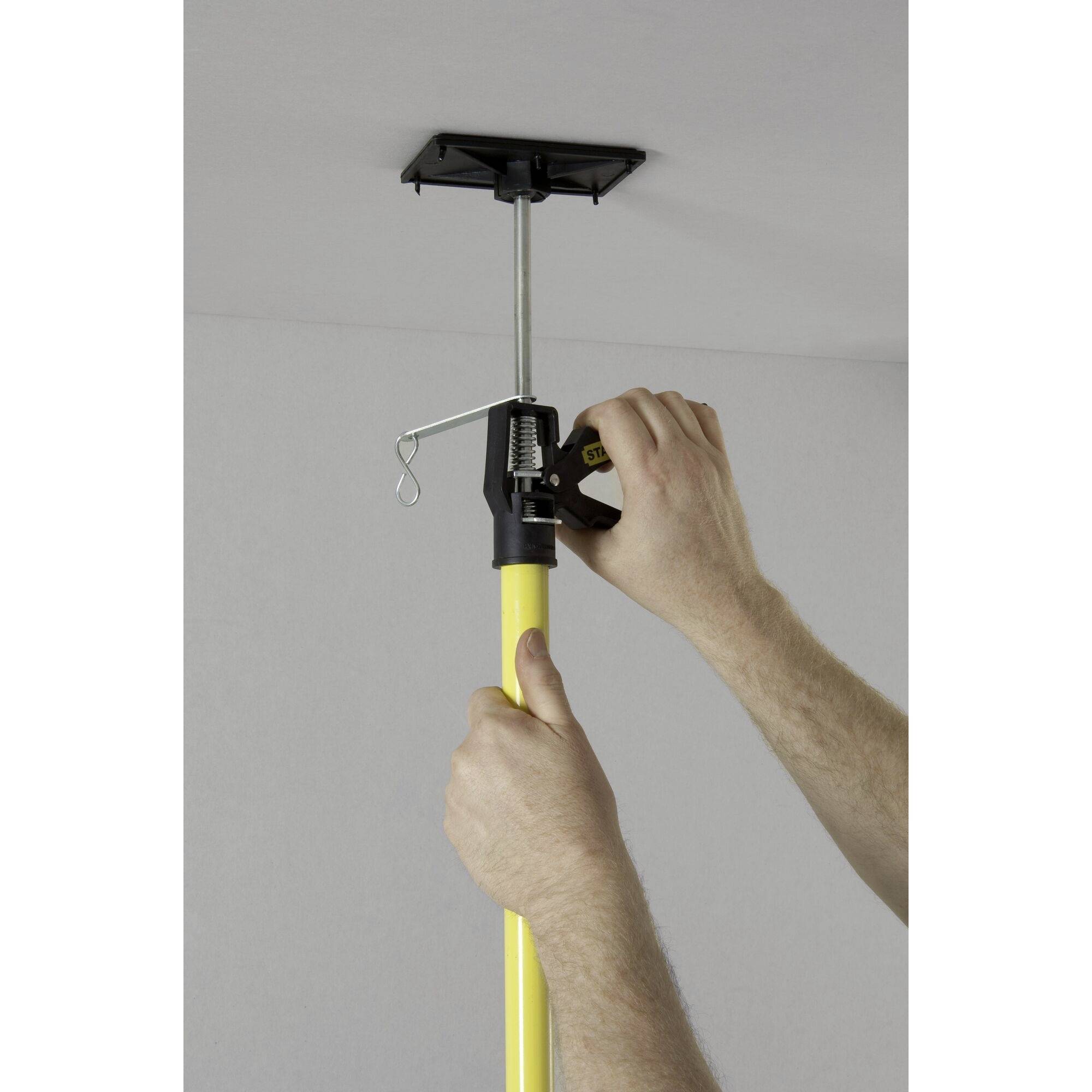 'A person attaches a yellow telescopic pole to the ceiling to secure a mounting bracket. Hands are turning the tensioning device.'