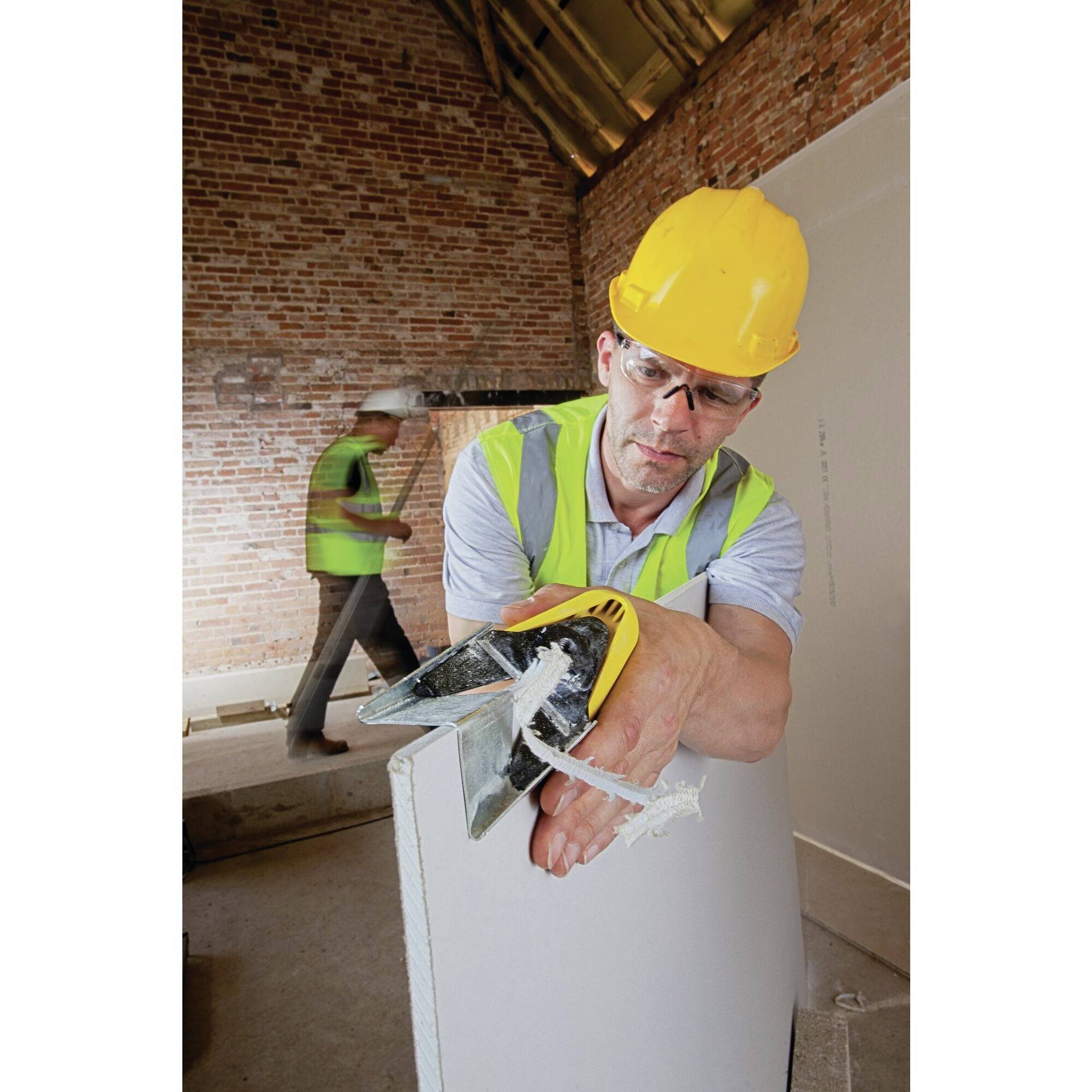A construction worker wearing a yellow hi-vis vest and hard hat is cutting a plasterboard. Another worker can be seen running in the background.