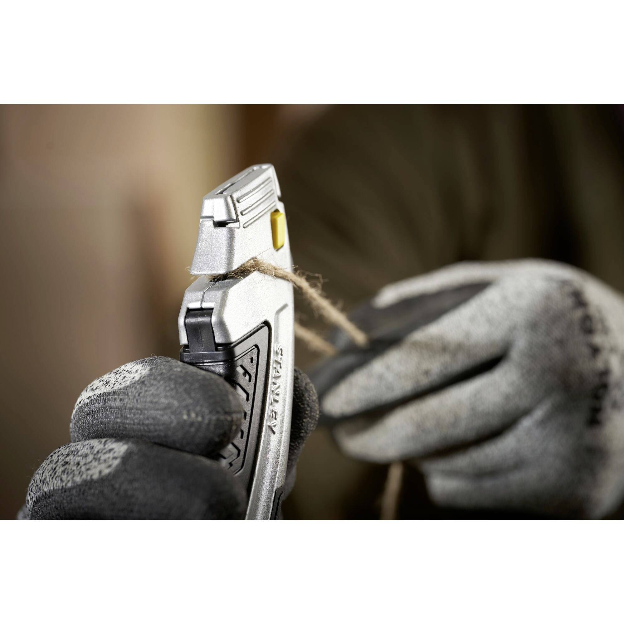 A person is cutting a thick rope with a silver craft knife. The hands are wearing grey work gloves.