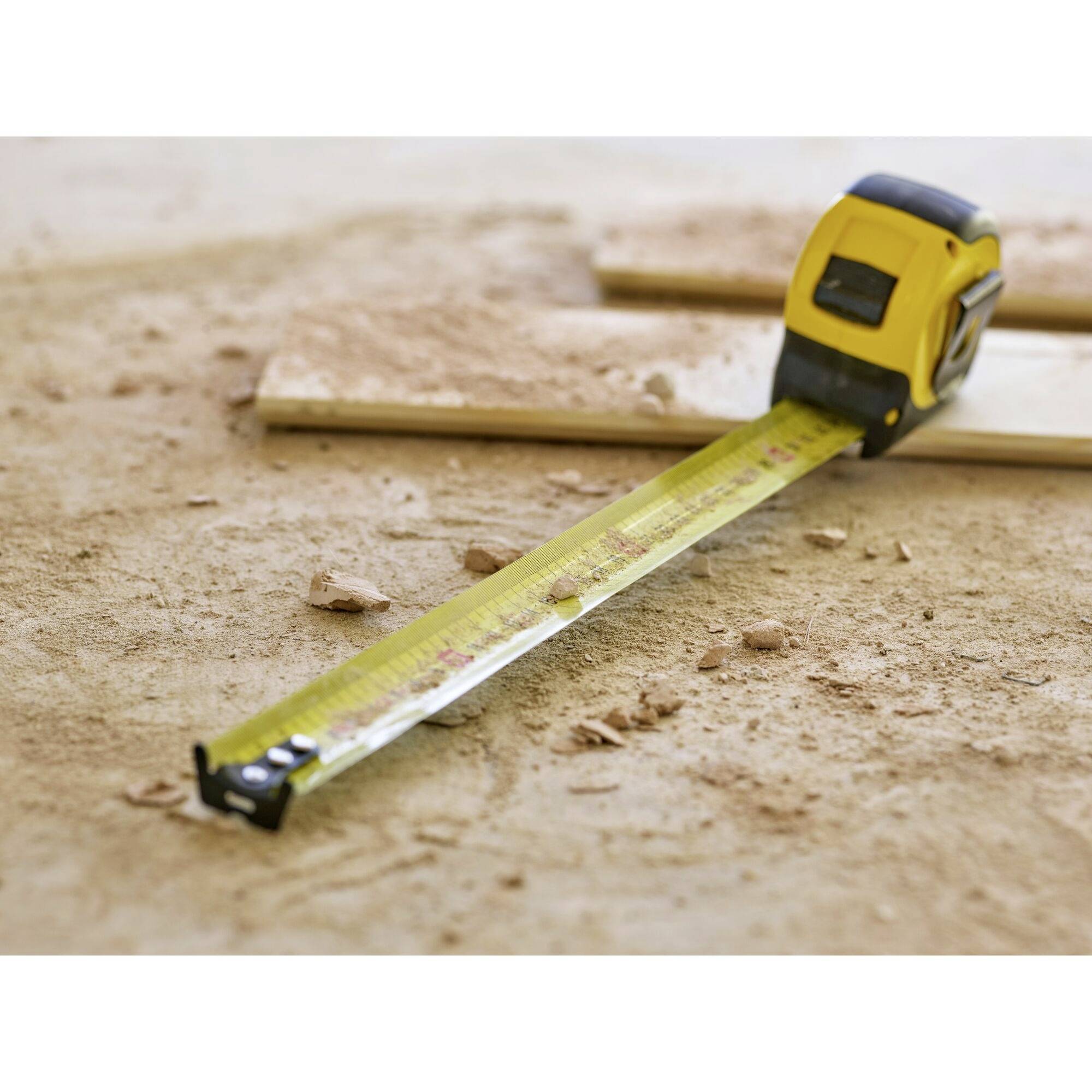 A yellow tape measure on dusty ground, stretched out in front of a wooden board. Signs of construction or renovation work.