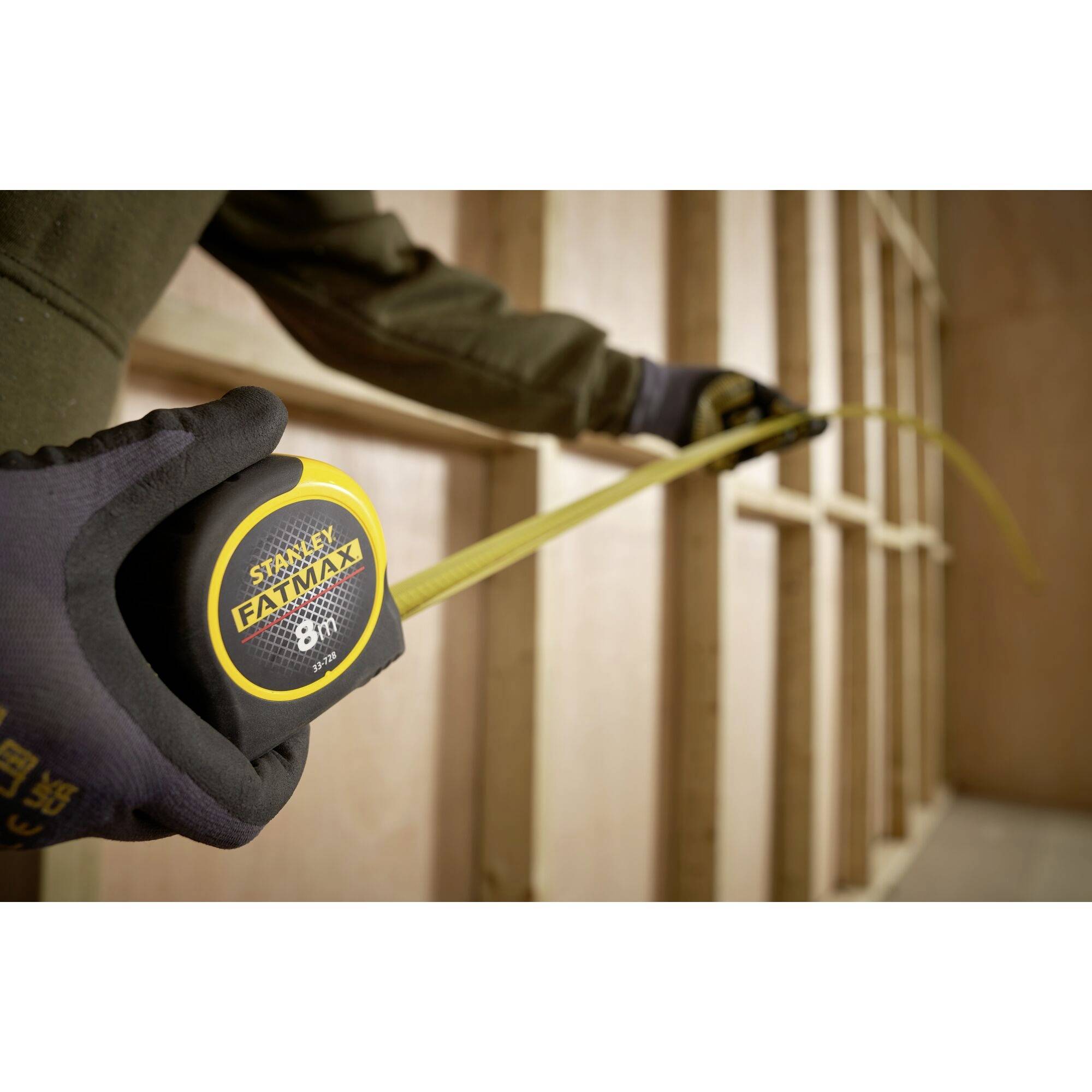 A person is measuring the distance between wooden frames using a 'Stanley FatMax 8m' tape measure. Hands wearing work gloves, with the focus on the measuring tape.