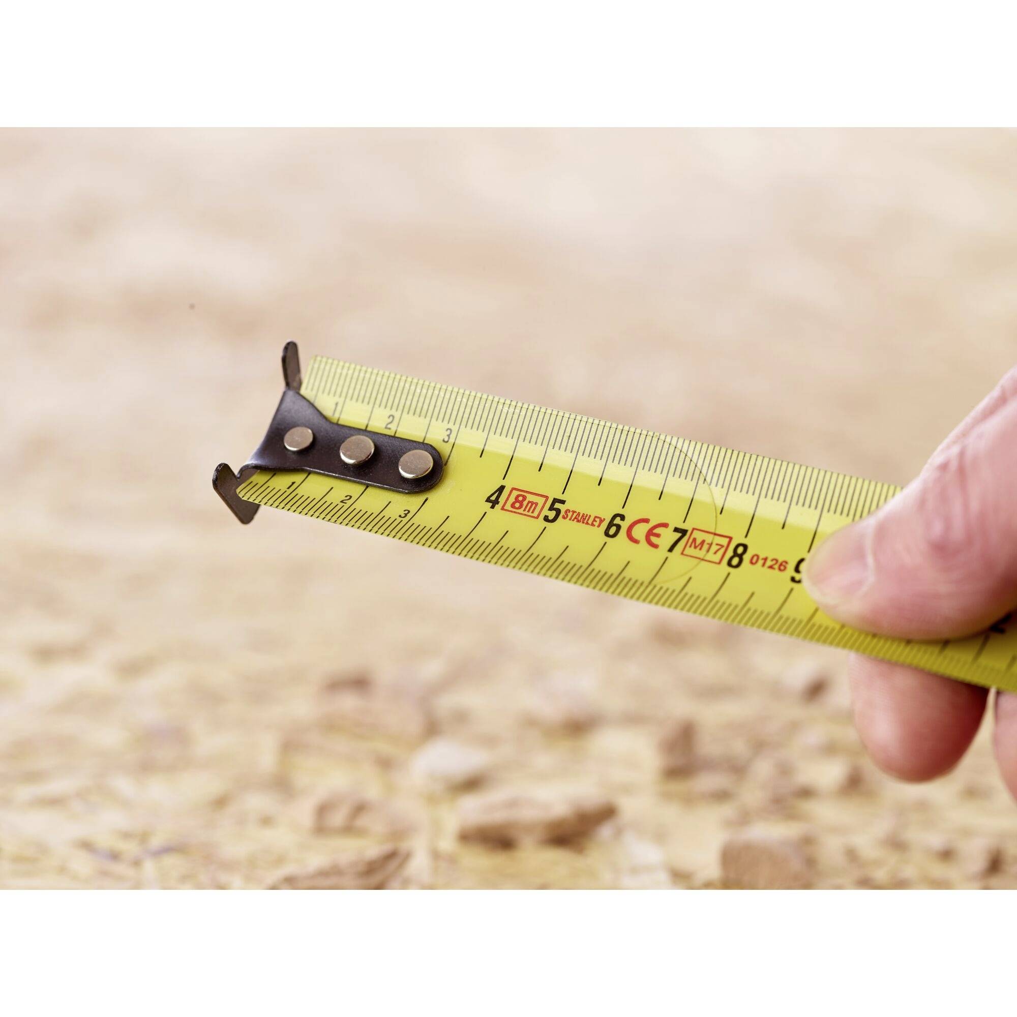 A tape measure is unrolled on an earthy ground. A hand is holding the tape, with its scale clearly visible.