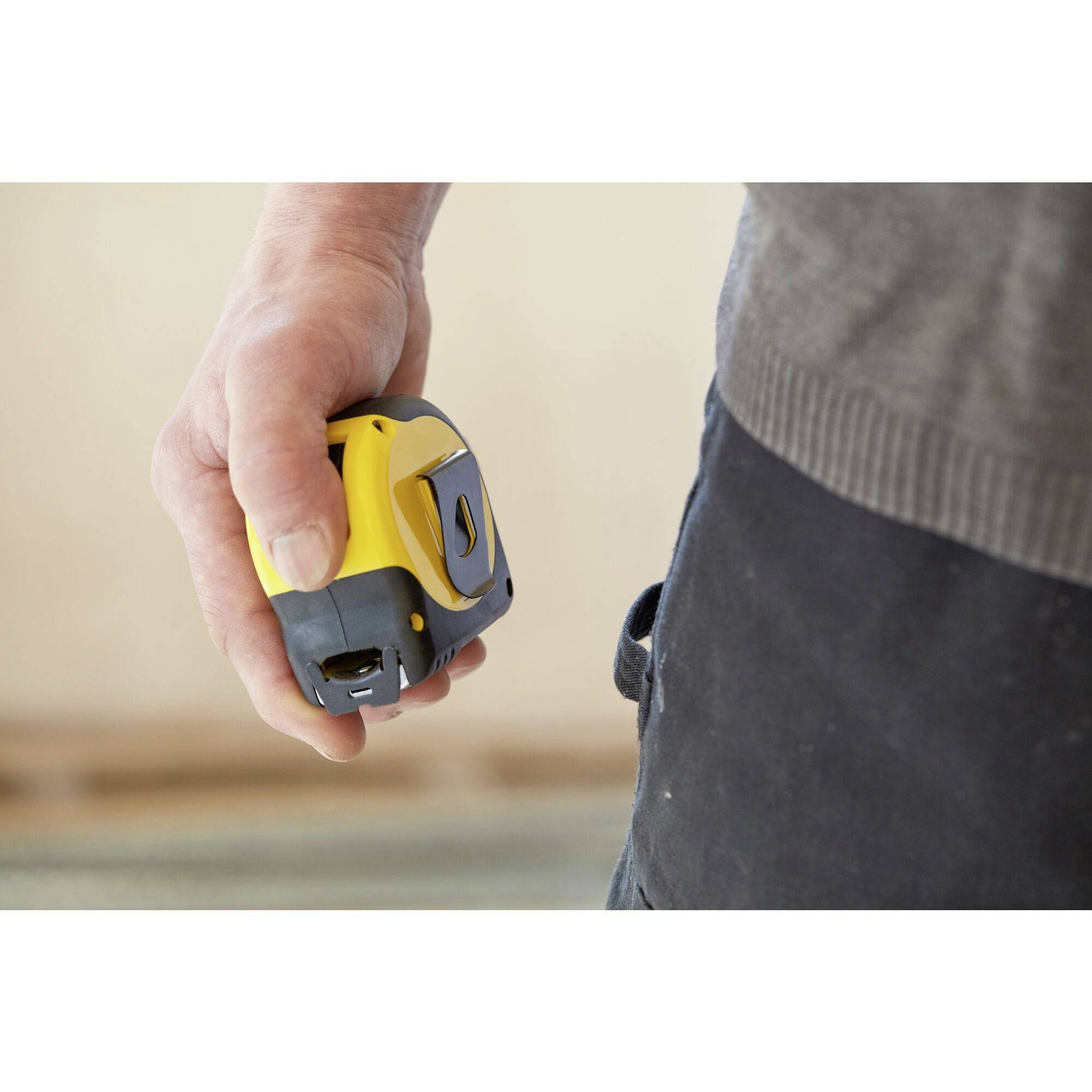 A person is holding a yellow tape measure in their hand, ready for use. A blurred interior is visible in the background.