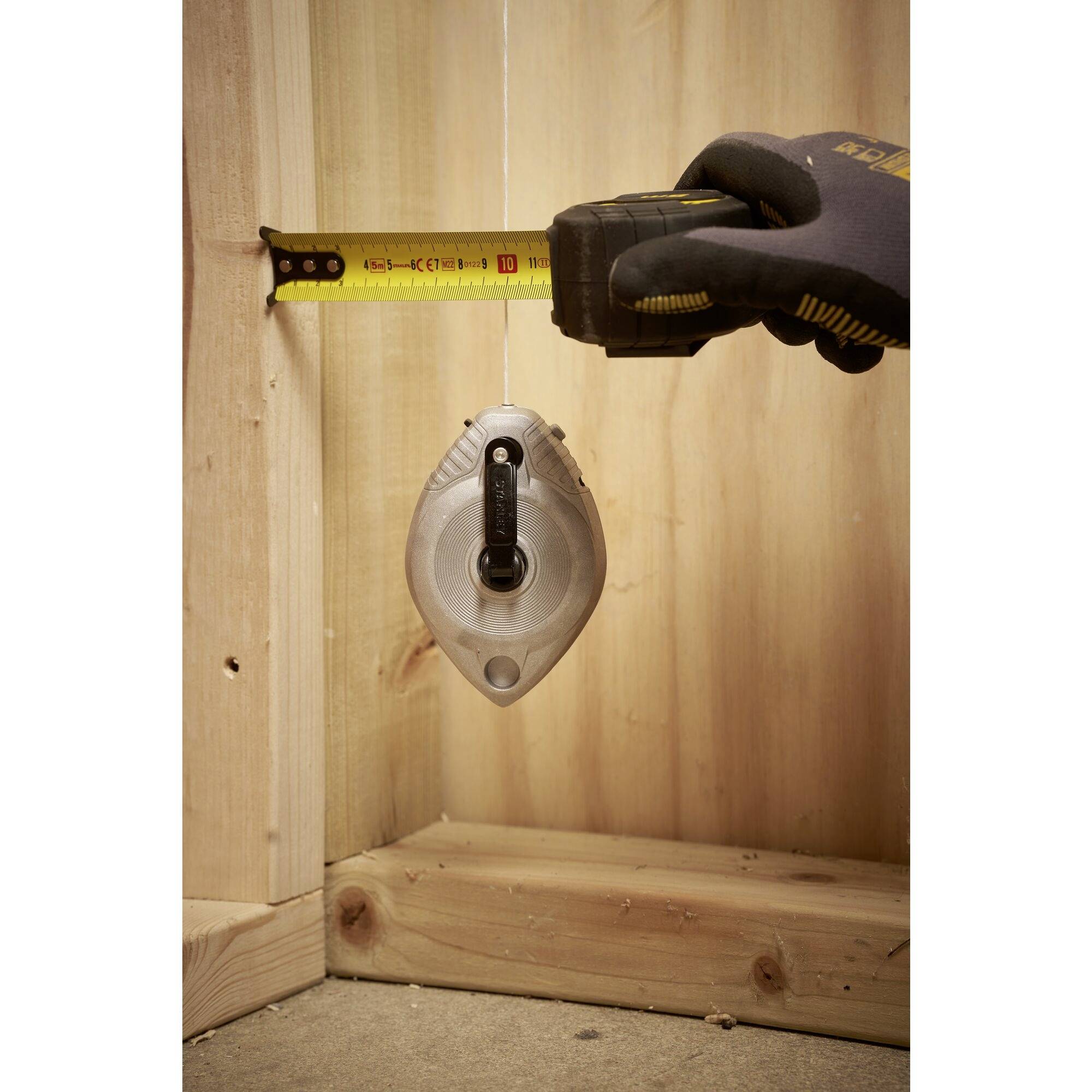 A hand is holding a tape measure against a wooden wall, next to a chalk marker. The setup is used for measuring and marking heights or lengths.