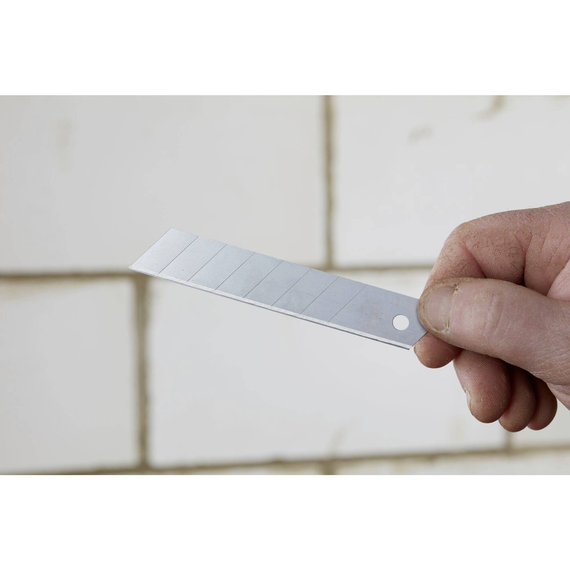 A hand holds a blade in front of a tiled wall, poised to cut or scrape.