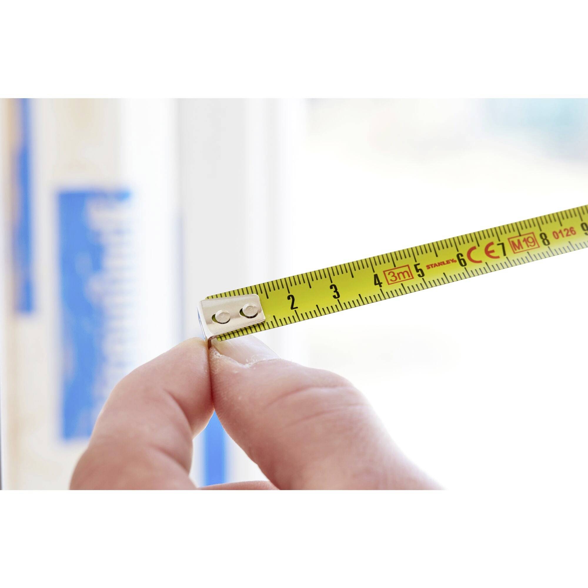 A hand holds a tape measure showing a length of 3 metres against a blurred background with blue and white elements.