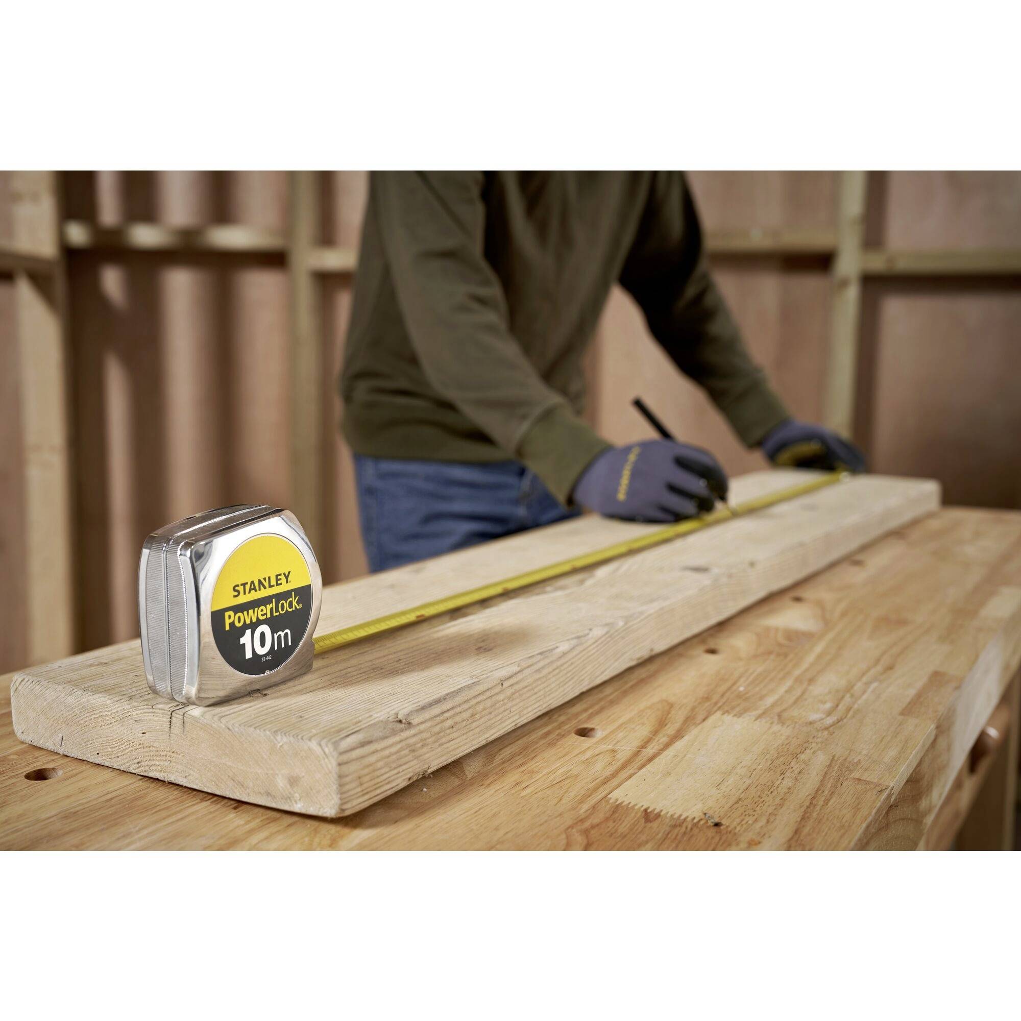 A person is measuring a long wooden board with a tape measure on a wooden table in a workshop, wearing gloves and noting down measurements.