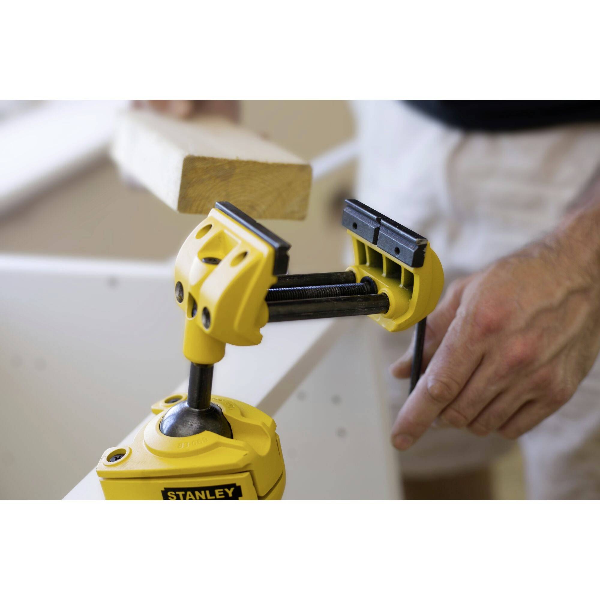 A yellow clamp holds a small piece of wood. A hand is adjusting the clamp. Workplace environment in the background.