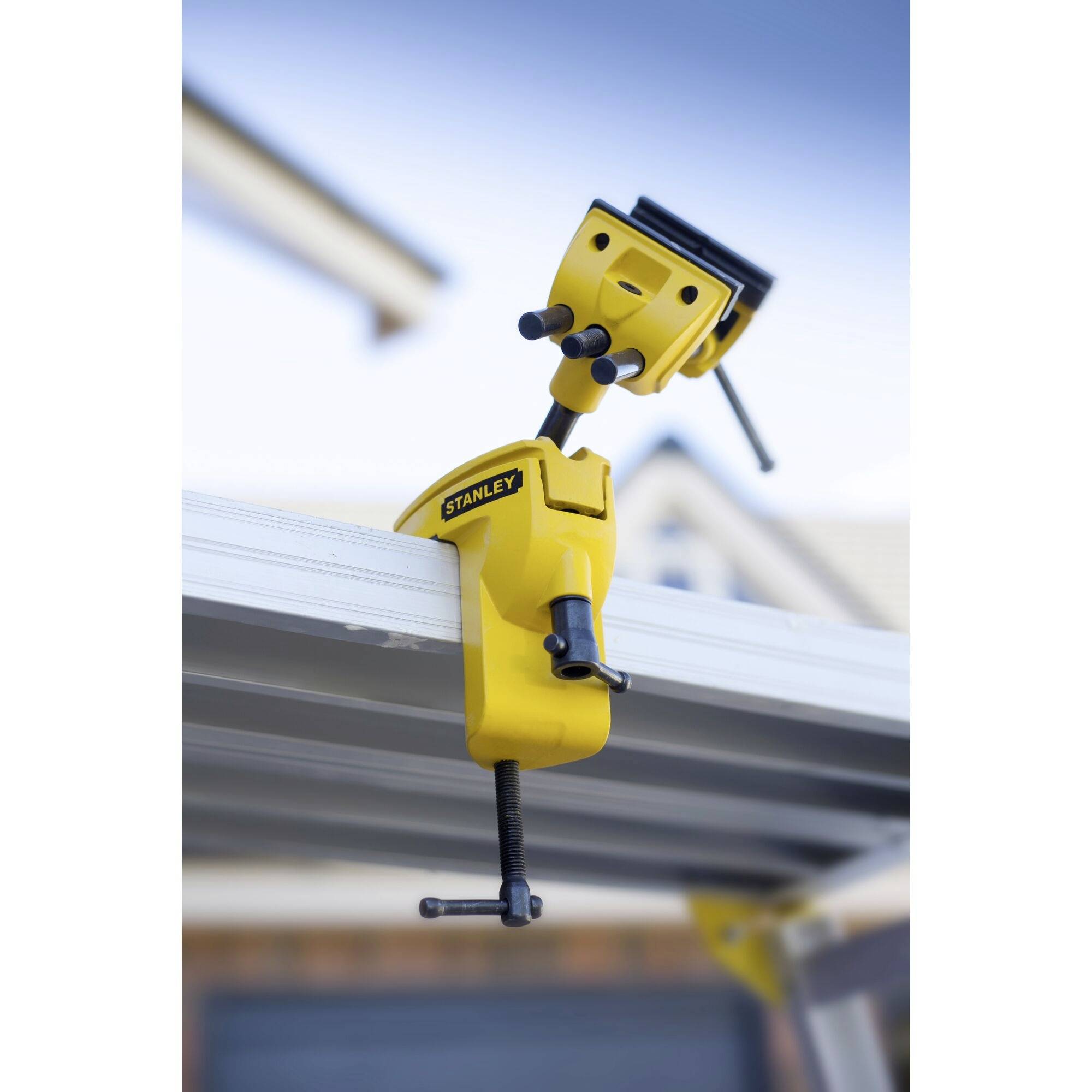 A yellow Stanley workbench G-clamp is secured to a metal surface. Blurred building outlines are visible in the background.