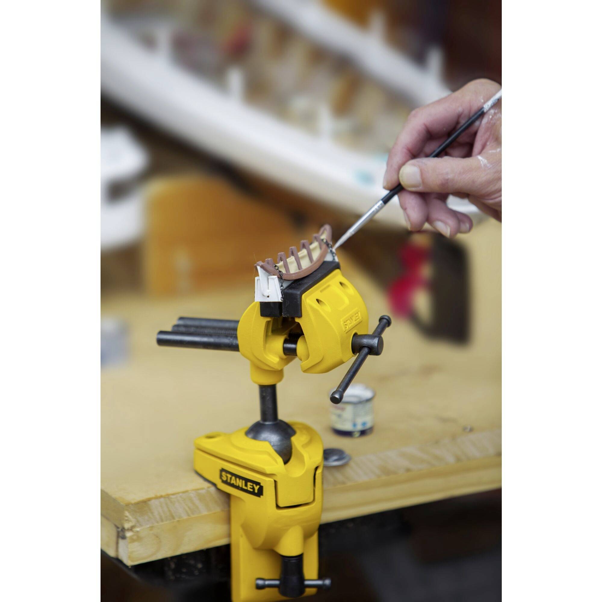A yellow G-clamp holds a model. A hand is painting the model with a brush. Tools are visible in the background.