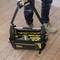 A person is carrying an open tool bag on a wooden floor. The bag is black and yellow and contains various tools.