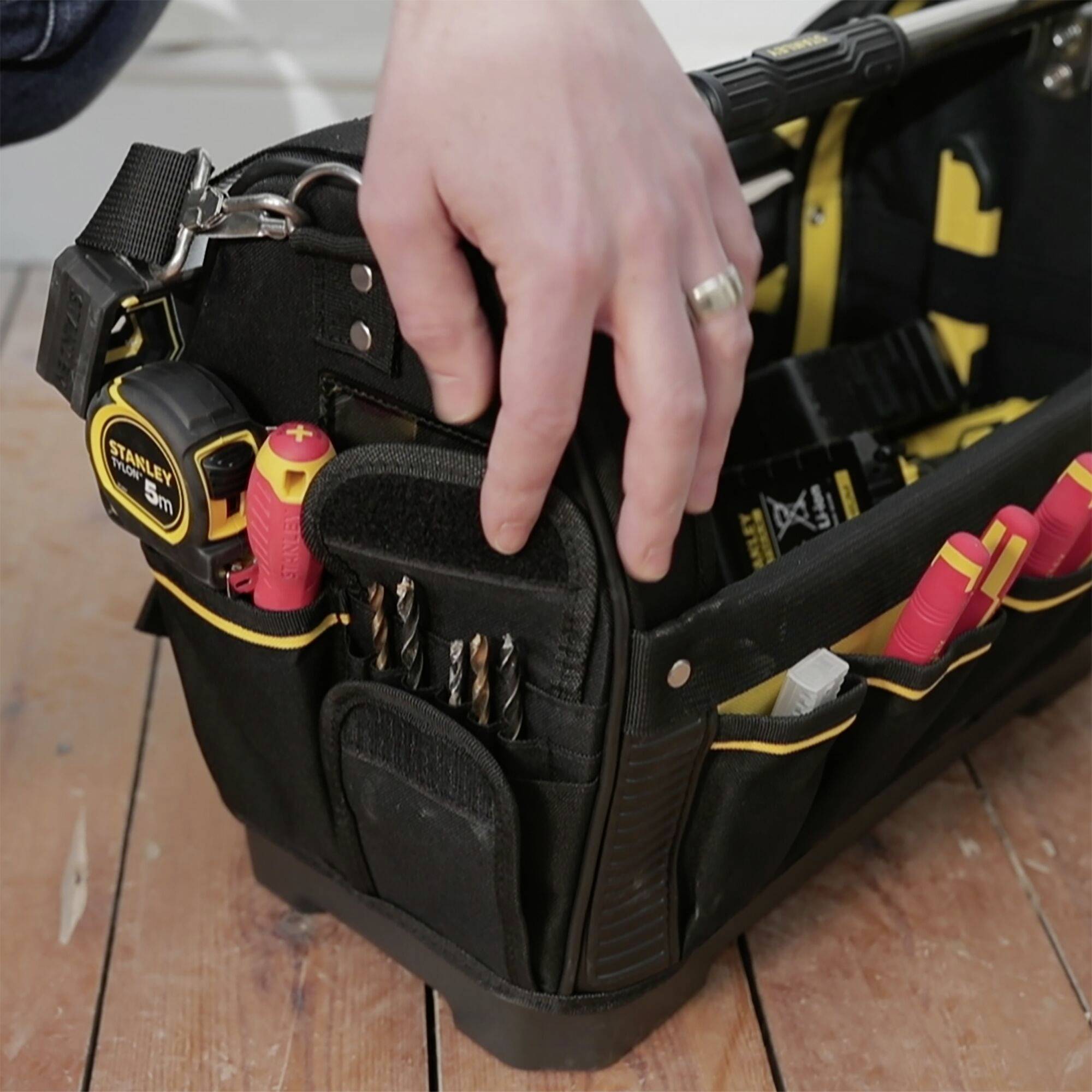 A hand reaches into an open tool bag containing various screwdrivers and tools, lying on a wooden floor.